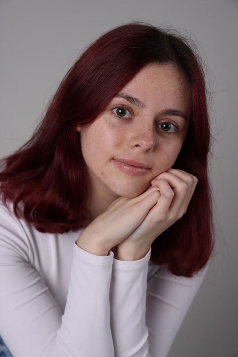 Young woman with red hair and a white shirt, resting her chin on her hands, gazing forward against a gray background.