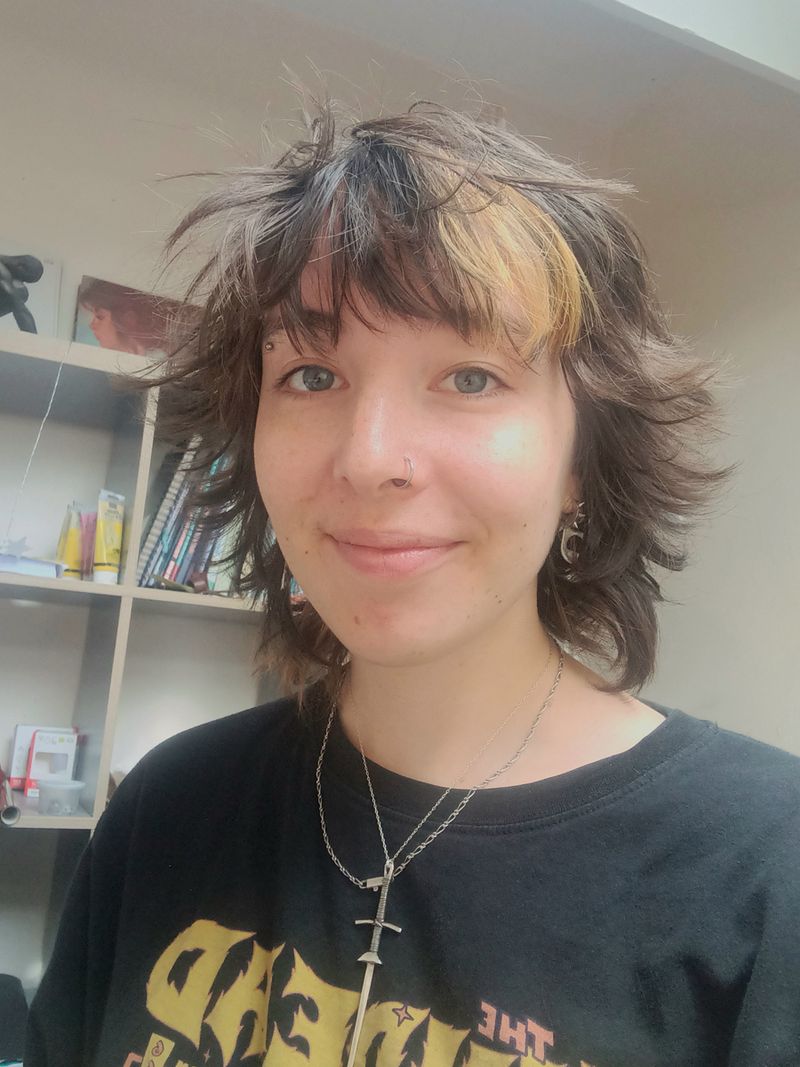 A person wearing a black graphic tee and a sword necklace smiles at the camera. A shelf with books is in the background.