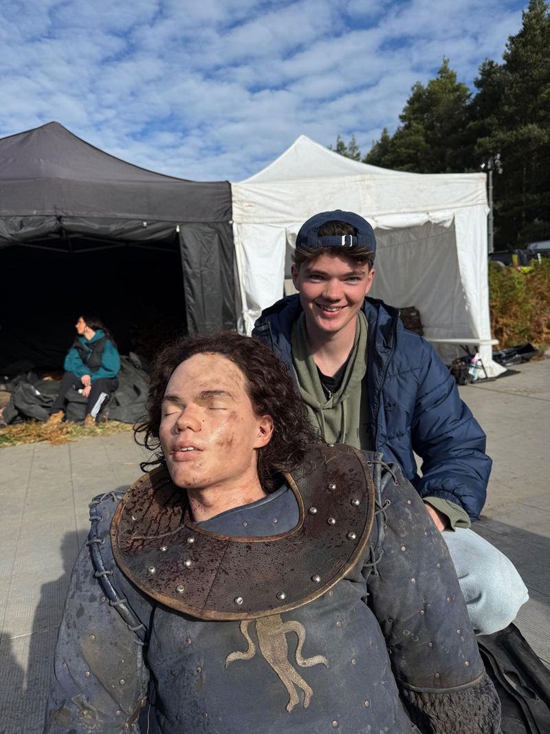 A person smiling behind a mannequin dressed in medieval armour, with tents and a cloudy sky in the background.