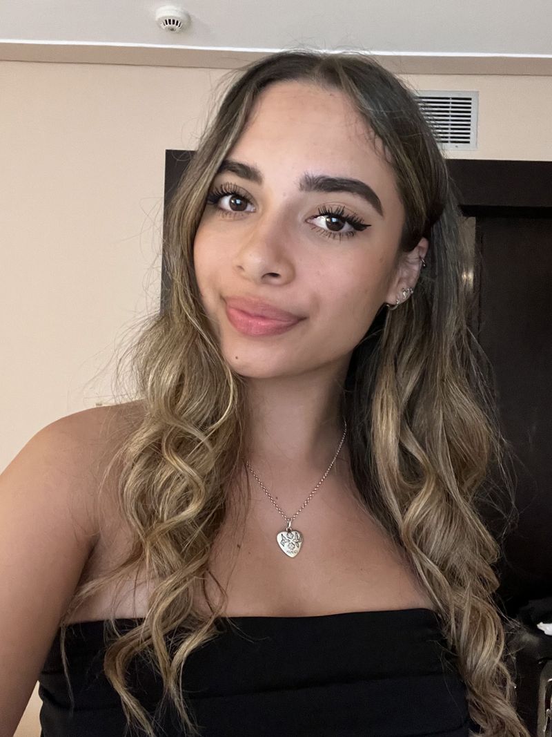 Headshot of Amelie Cooke. Amelie has long wavy blonde hair, wearing a strapless top and heart necklace. Amelie smiles at the camera in an indoor setting.
