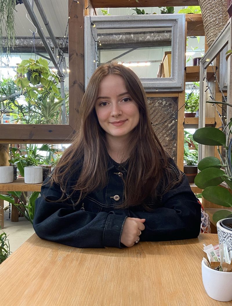 Ruby Brown with long brown hair sits at a wooden table in a plant-filled setting, wearing a black jacket, and smiling at the camera.