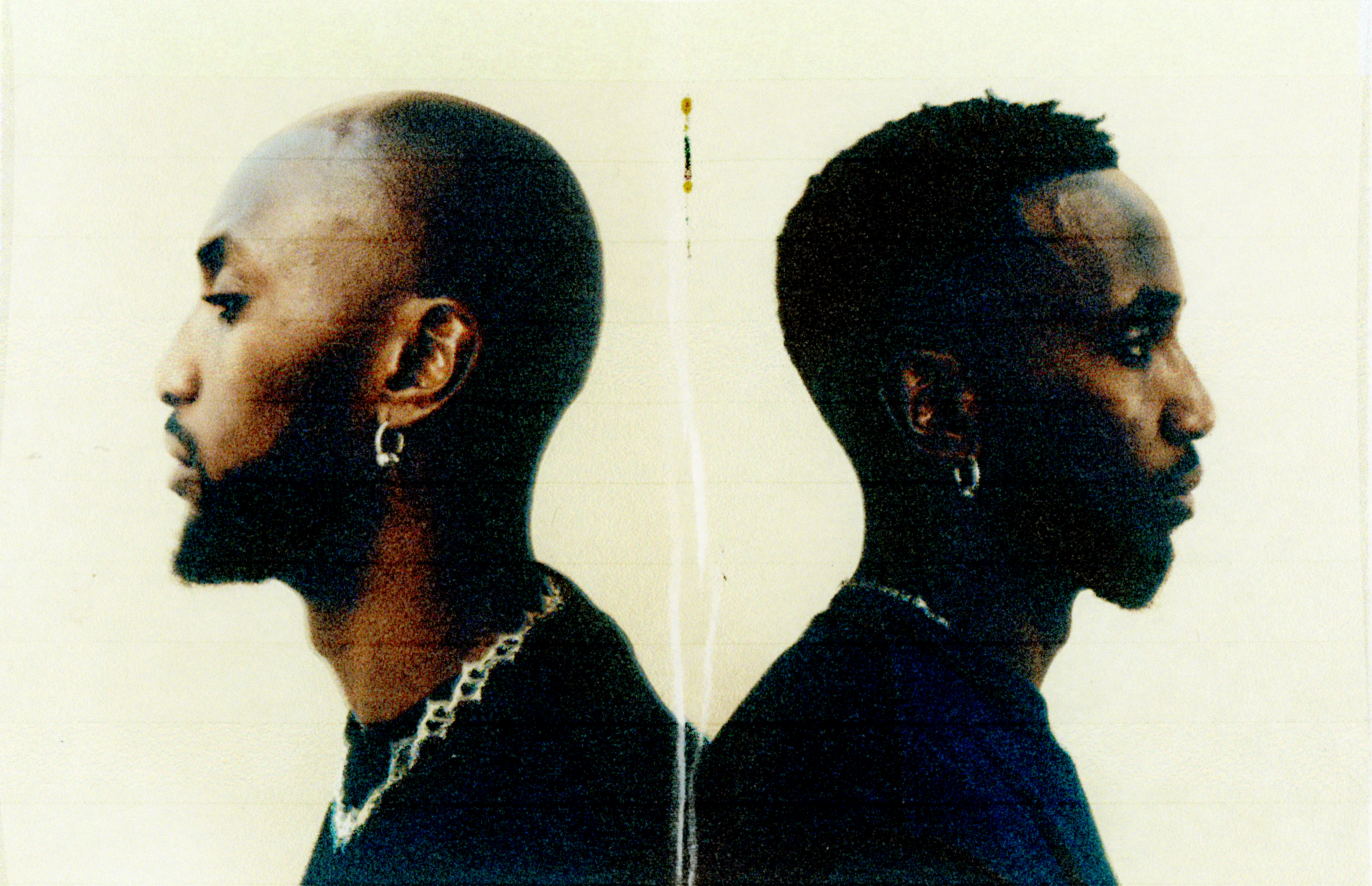 Deki Alem standing back-to-back, facing opposite directions. Both wear earrings and dark clothing against a light background.