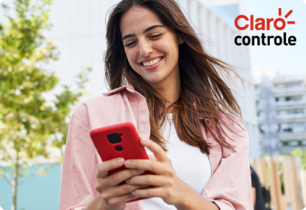 Mulher sorrir e mexe em um smartphone vermelho que está em suas mãos, veste blusa branca e jaqueta rosa. Foto ao ar livre com a logo do "Claro controle" no canto superior direito.