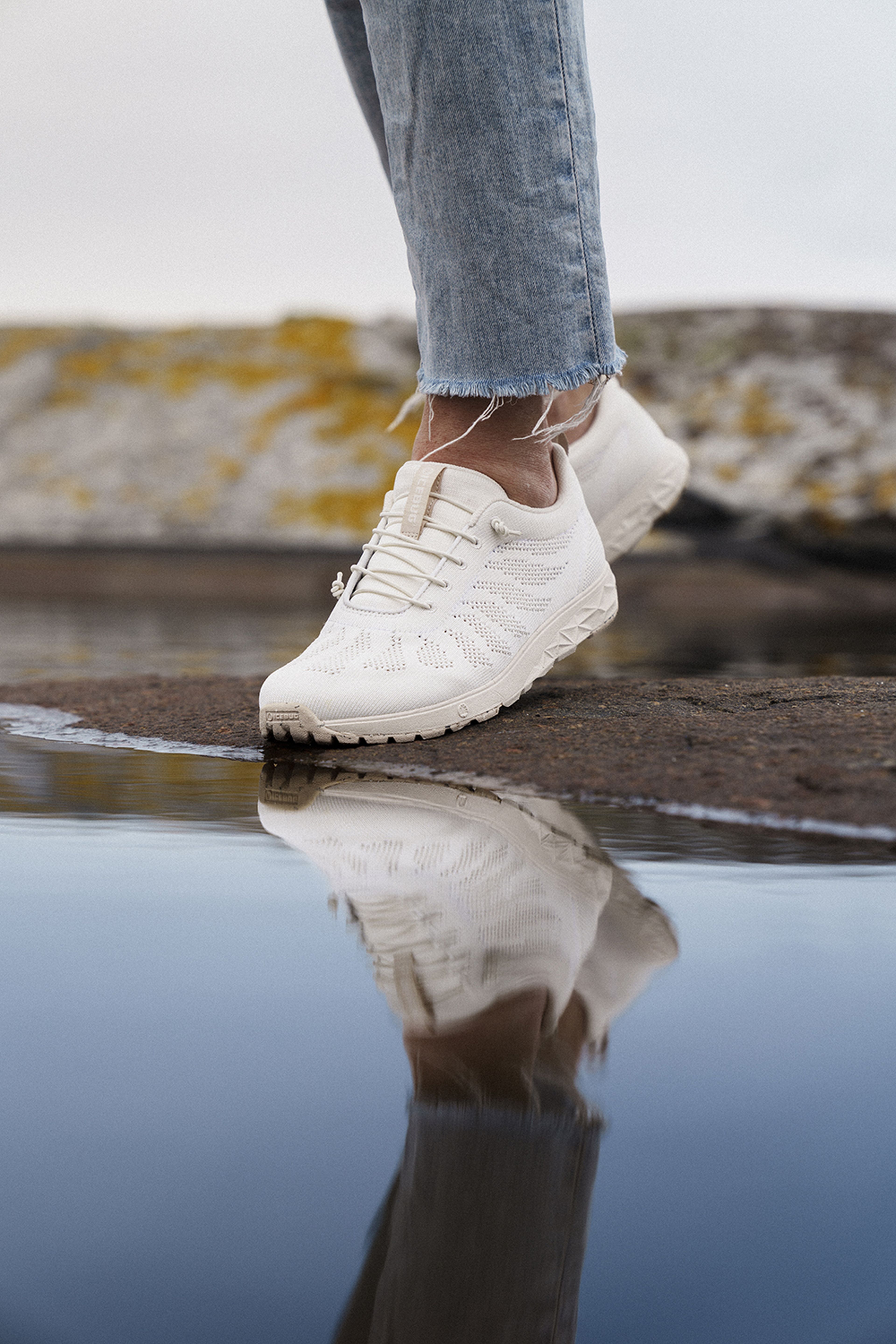 Person wearing white sneakers and frayed jeans stands on wet ground, with the shoe's reflection visible in the water.