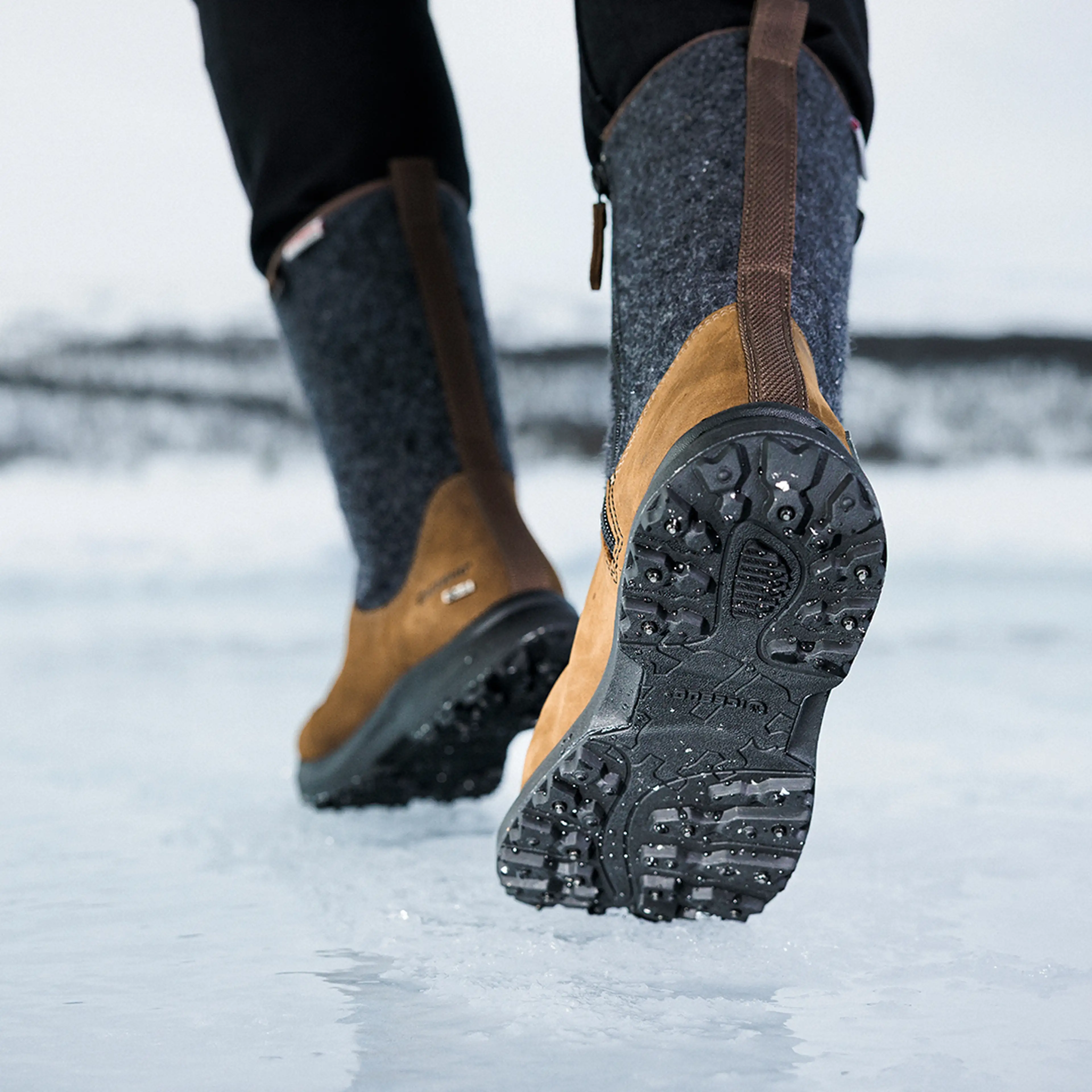 Close-up of a person walking on ice, wearing brown winter boots with studded soles, set in a snowy landscape.