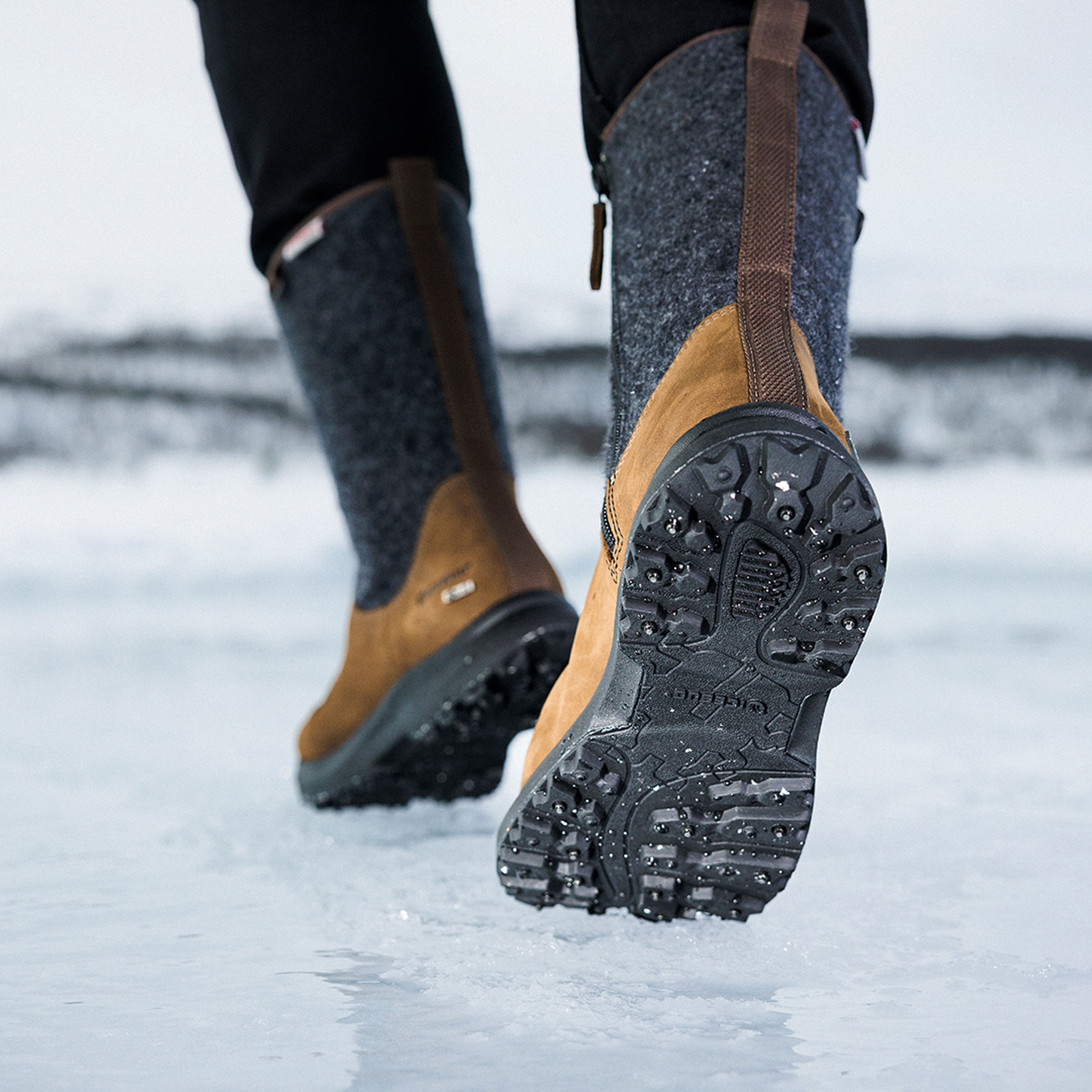 Close-up of a person walking on ice, wearing brown winter boots with studded soles, set in a snowy landscape.