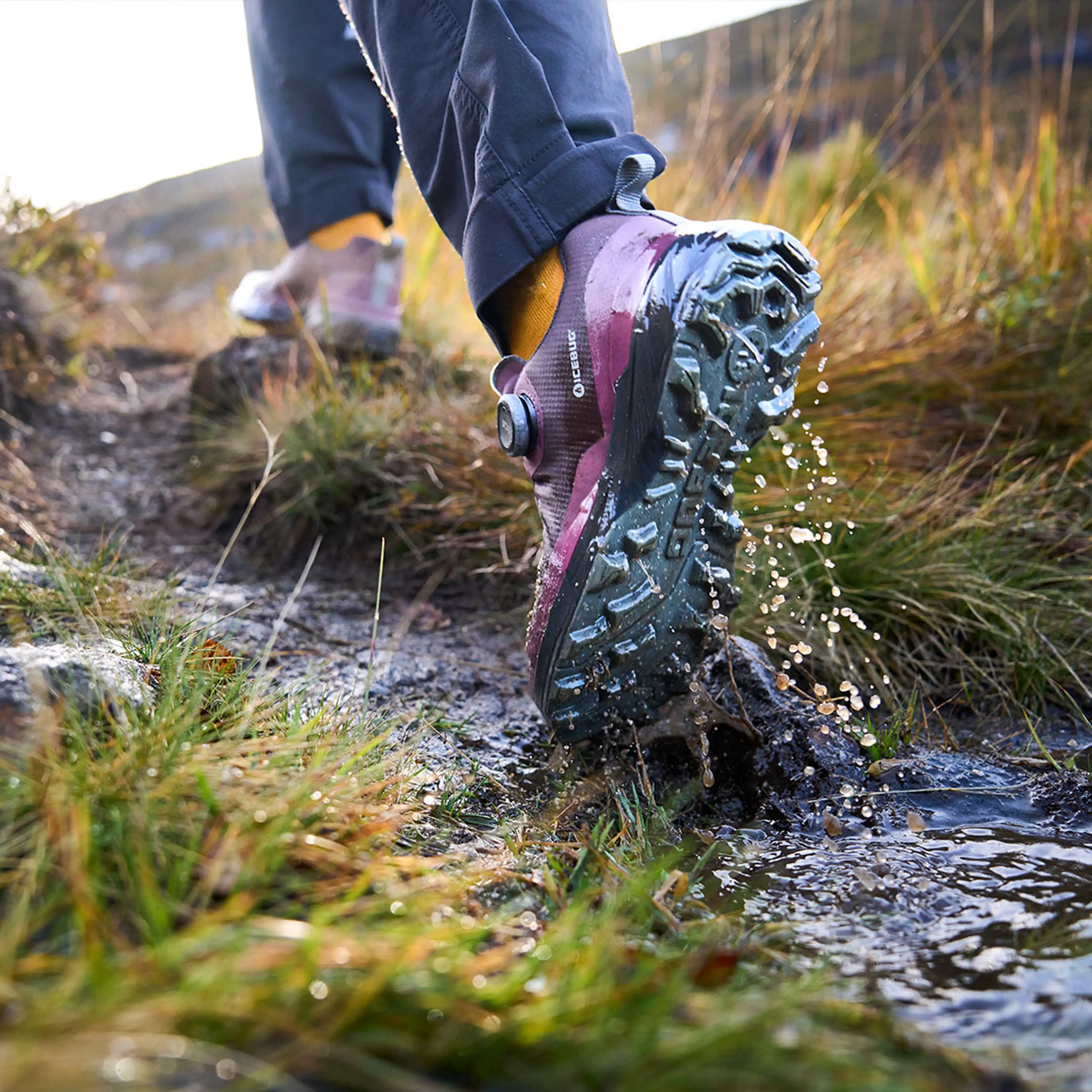 Person wearing purple hiking shoes steps in a muddy puddle on a trail, with water splashing, surrounded by green grass and hilly terrain.