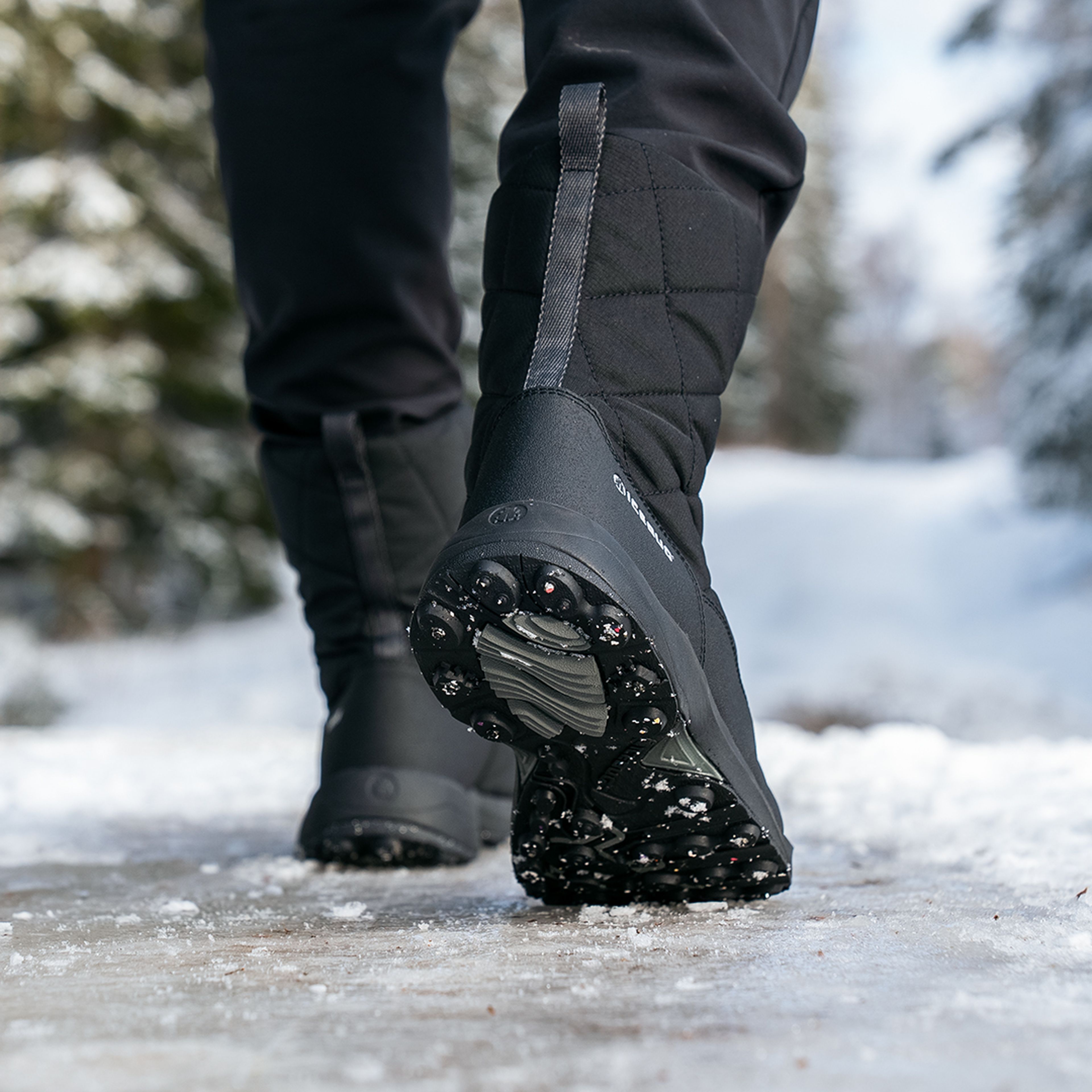 Close-up of black quilted winter boots on a snowy path, surrounded by tall trees.