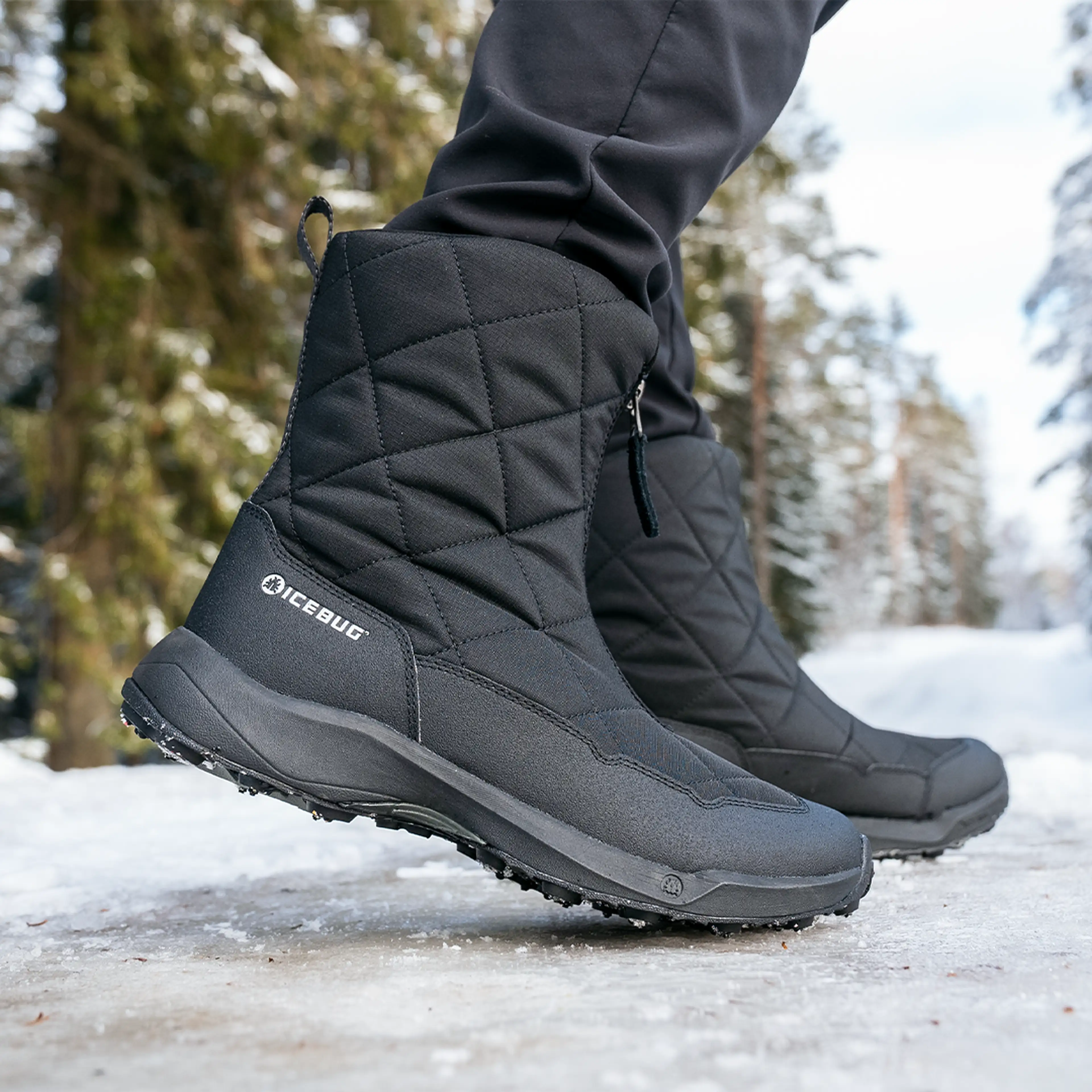 Close-up of black quilted winter boots on a snowy path, surrounded by tall evergreen trees.
