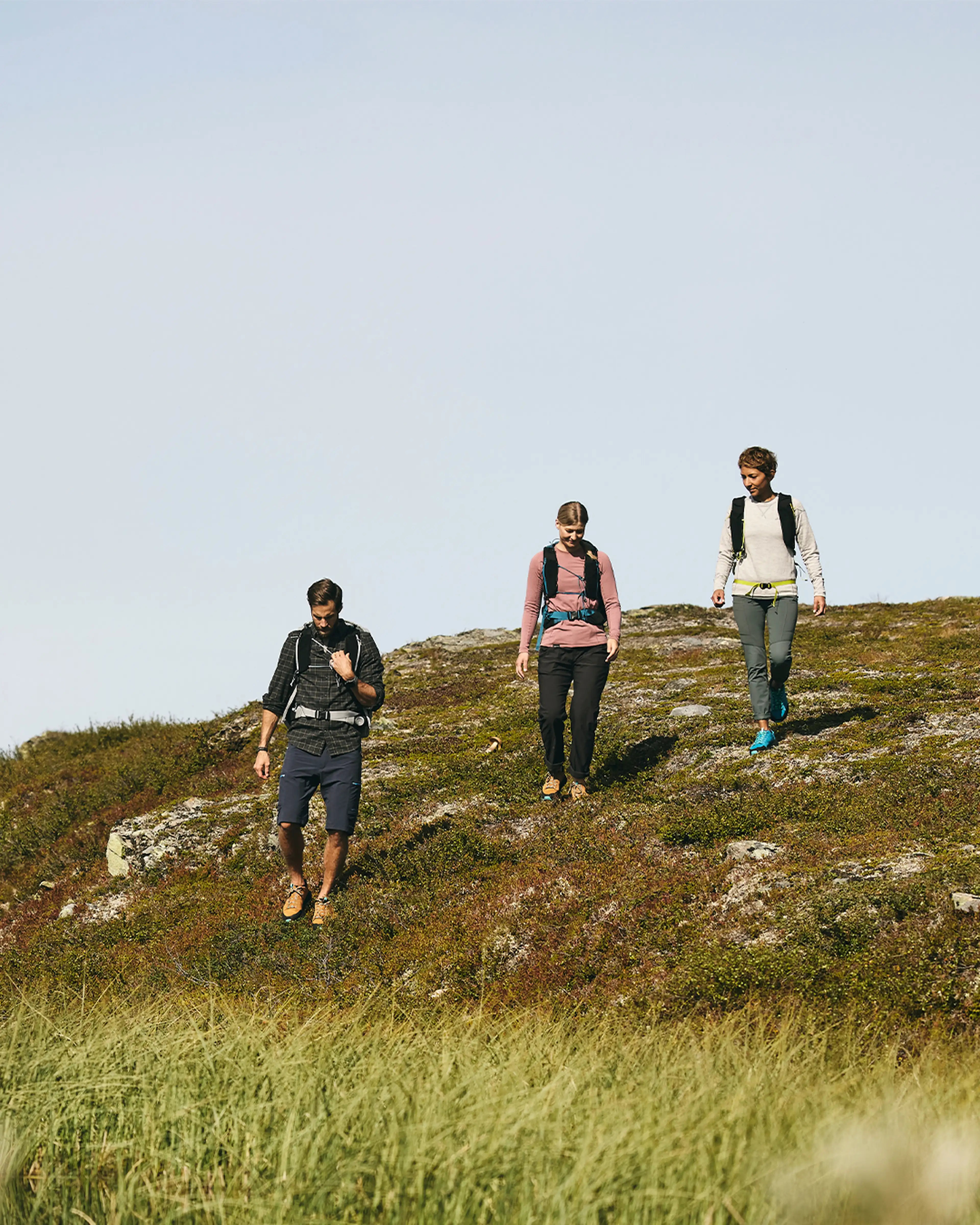 Three people hiking down a grassy hill under a clear sky, wearing backpacks and outdoor clothing.