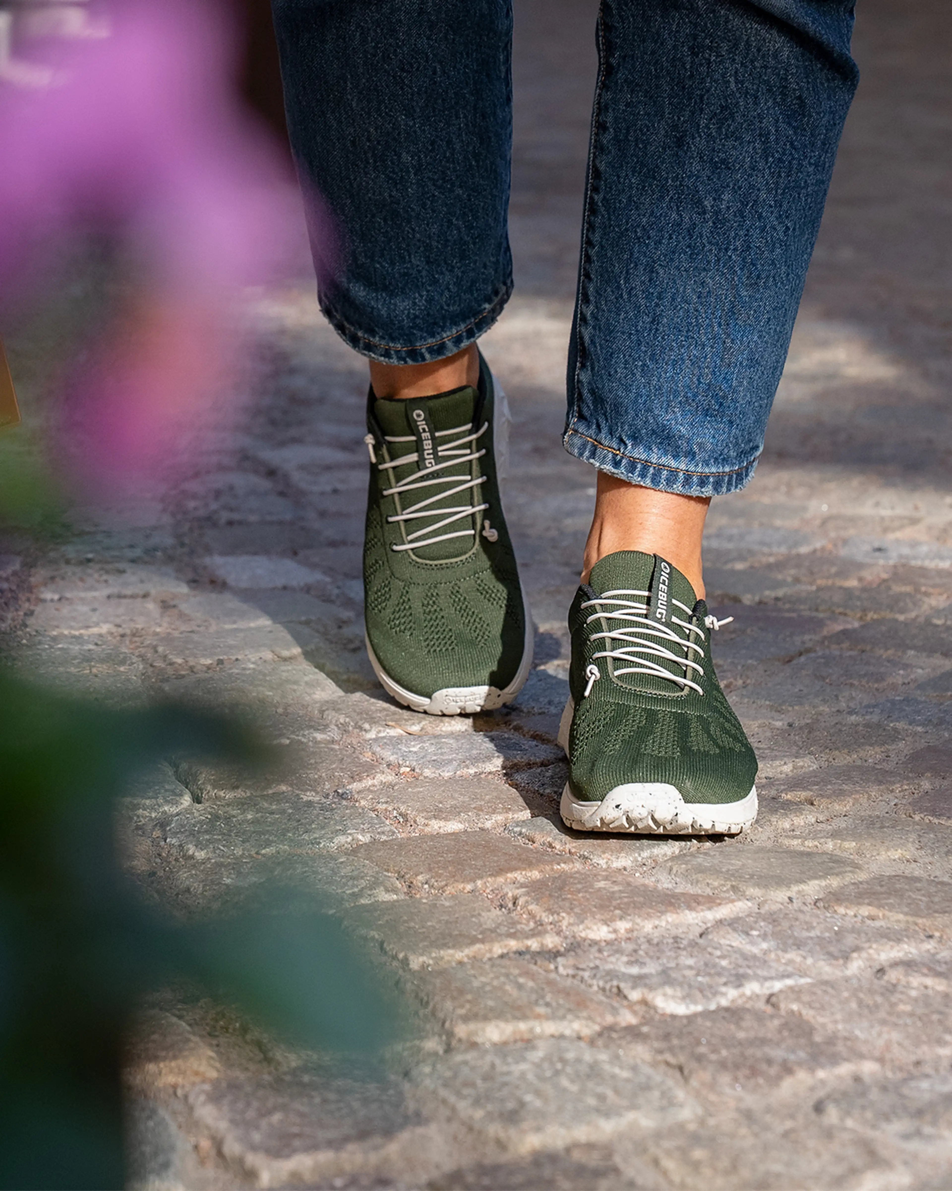 Person wearing green sneakers and jeans walking on a cobblestone path, with blurred greenery in the foreground.