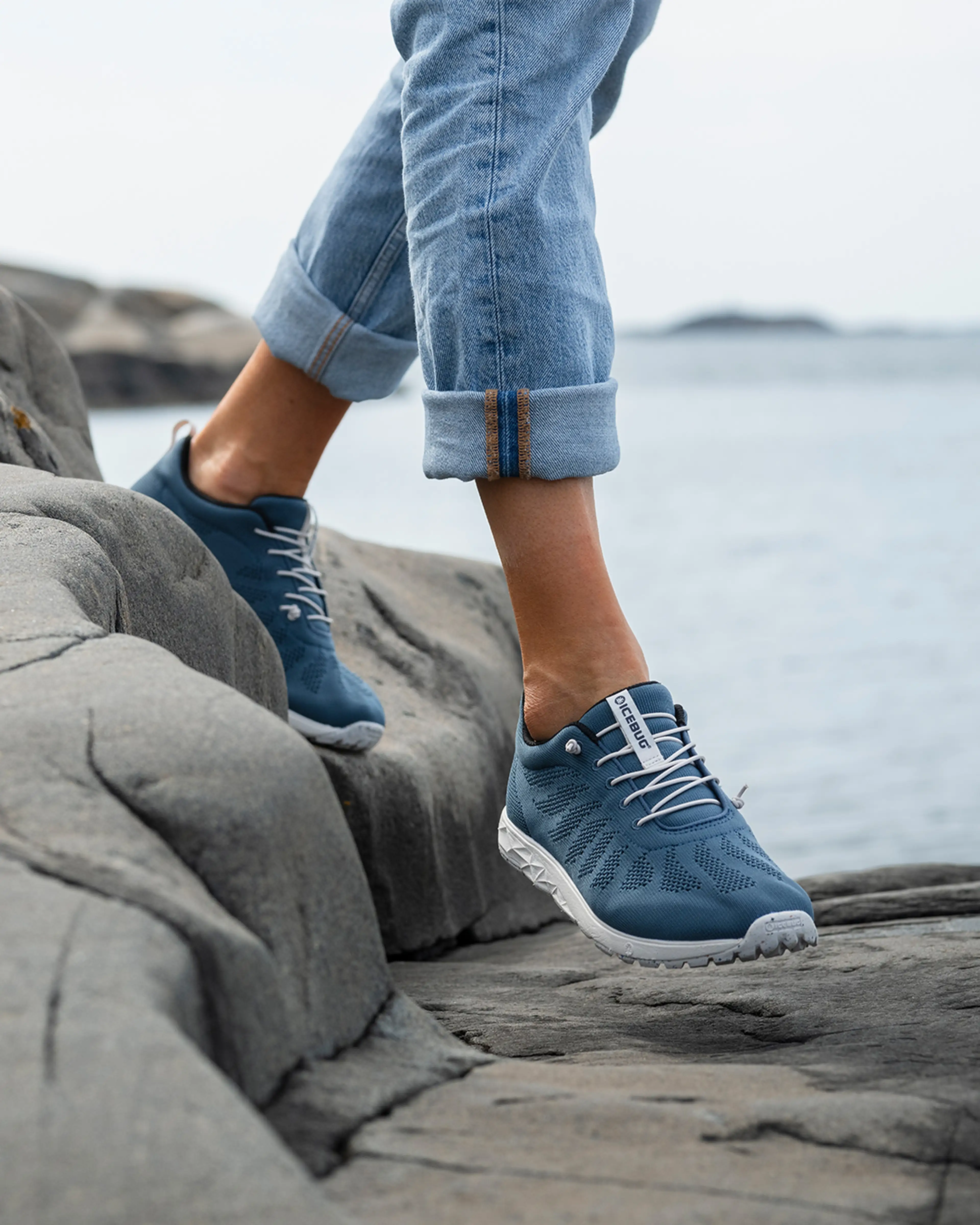 Person walking on rocky terrain near the water, wearing rolled-up jeans and blue sneakers with white soles.
