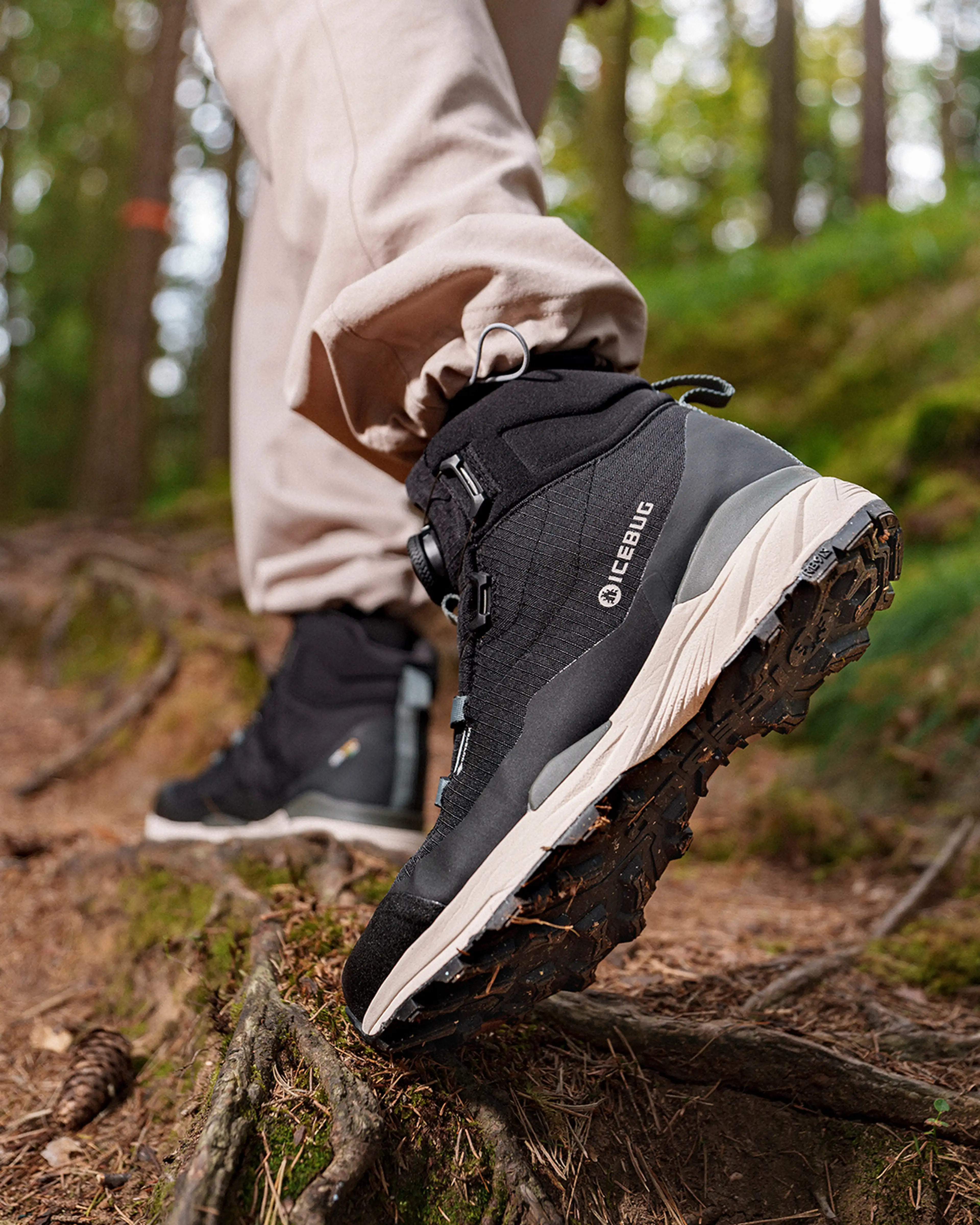 Close-up of a person hiking in a forest, wearing black Icebug boots and beige pants, stepping over tree roots on a dirt path.