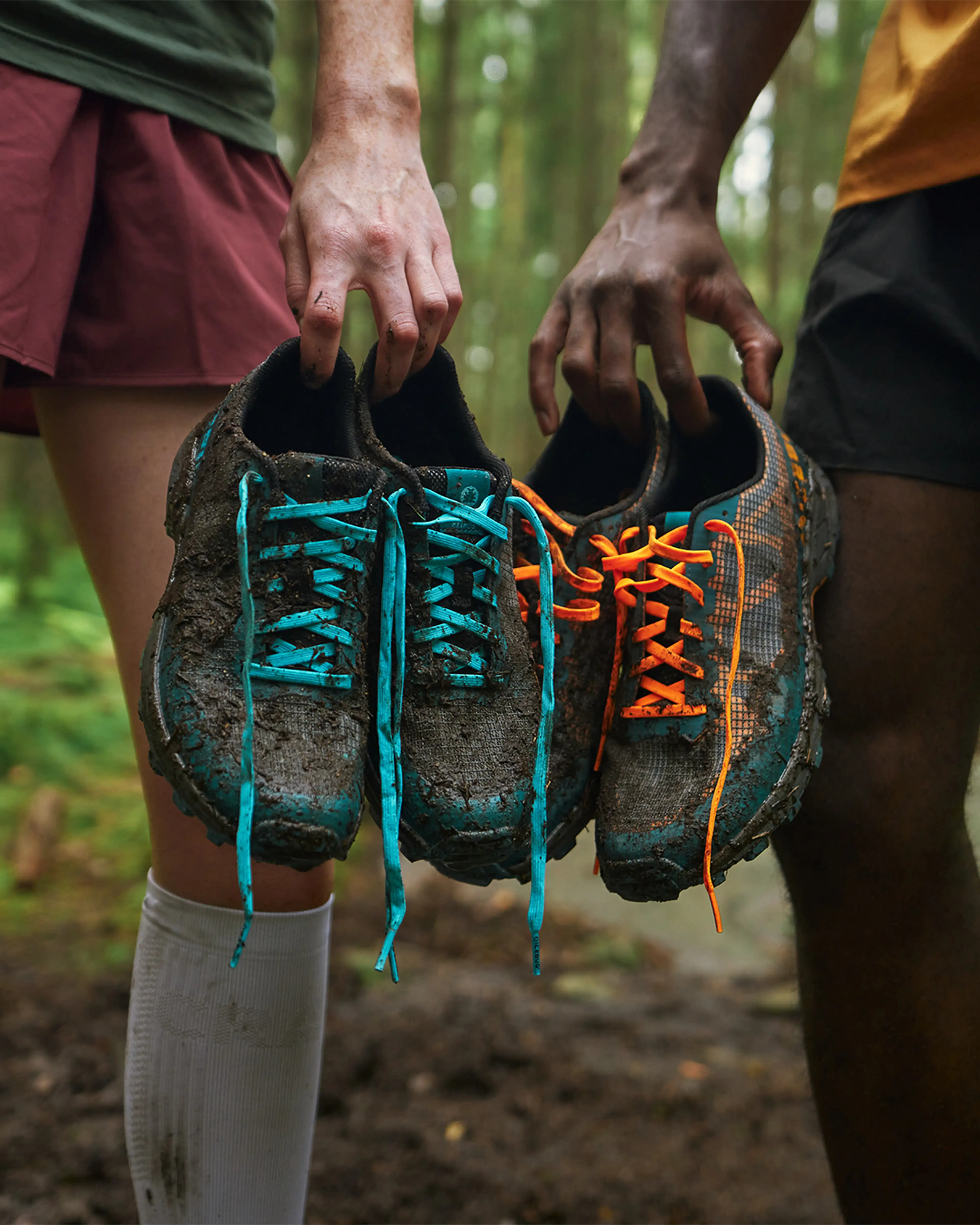 Two people holding muddy trail running shoes in a forest. One pair has blue laces, the other orange. Their legs are visible, wearing shorts.