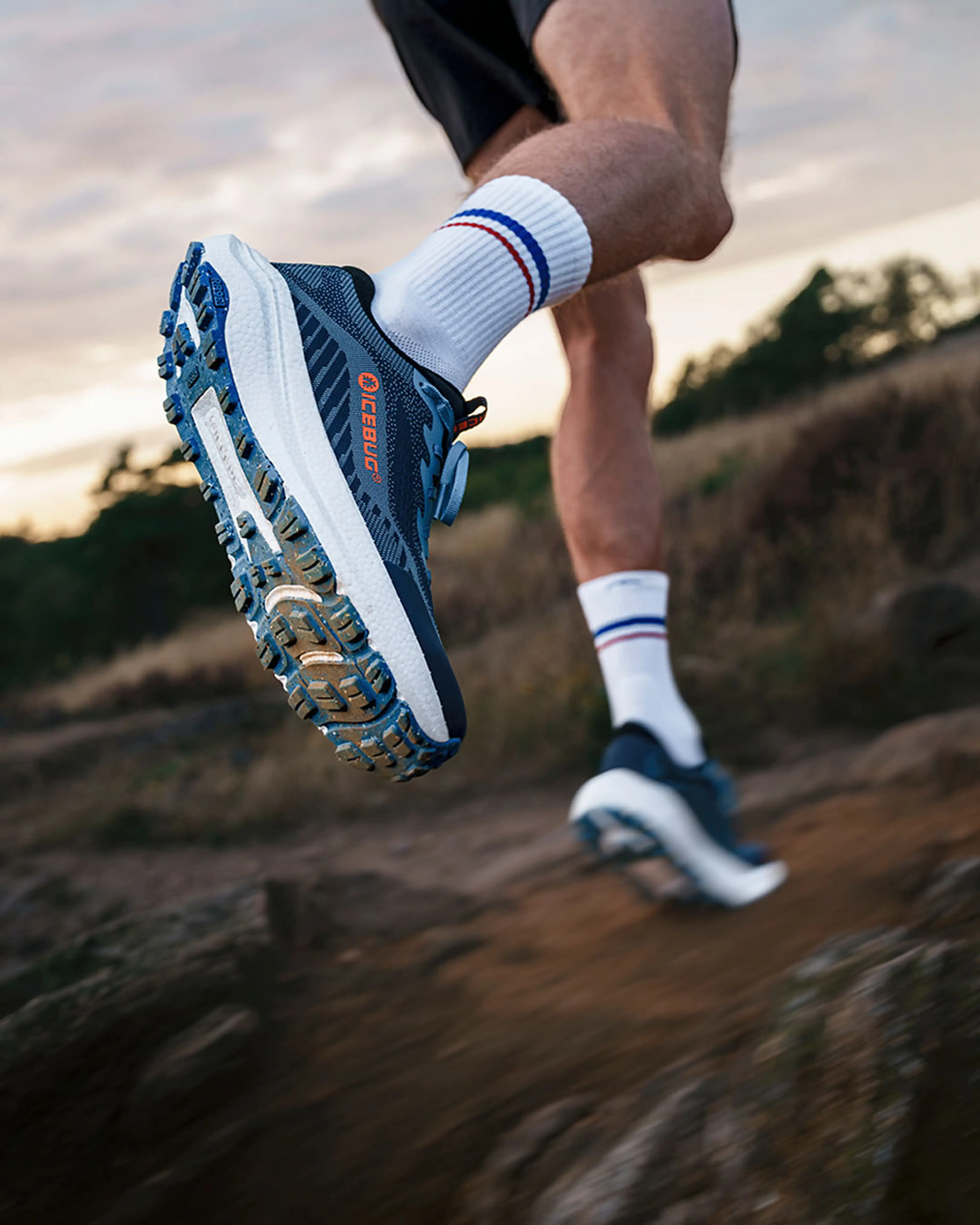 Close-up of a person running on a trail, focusing on the blue and white treaded sneaker with a dynamic, low-angle perspective.
