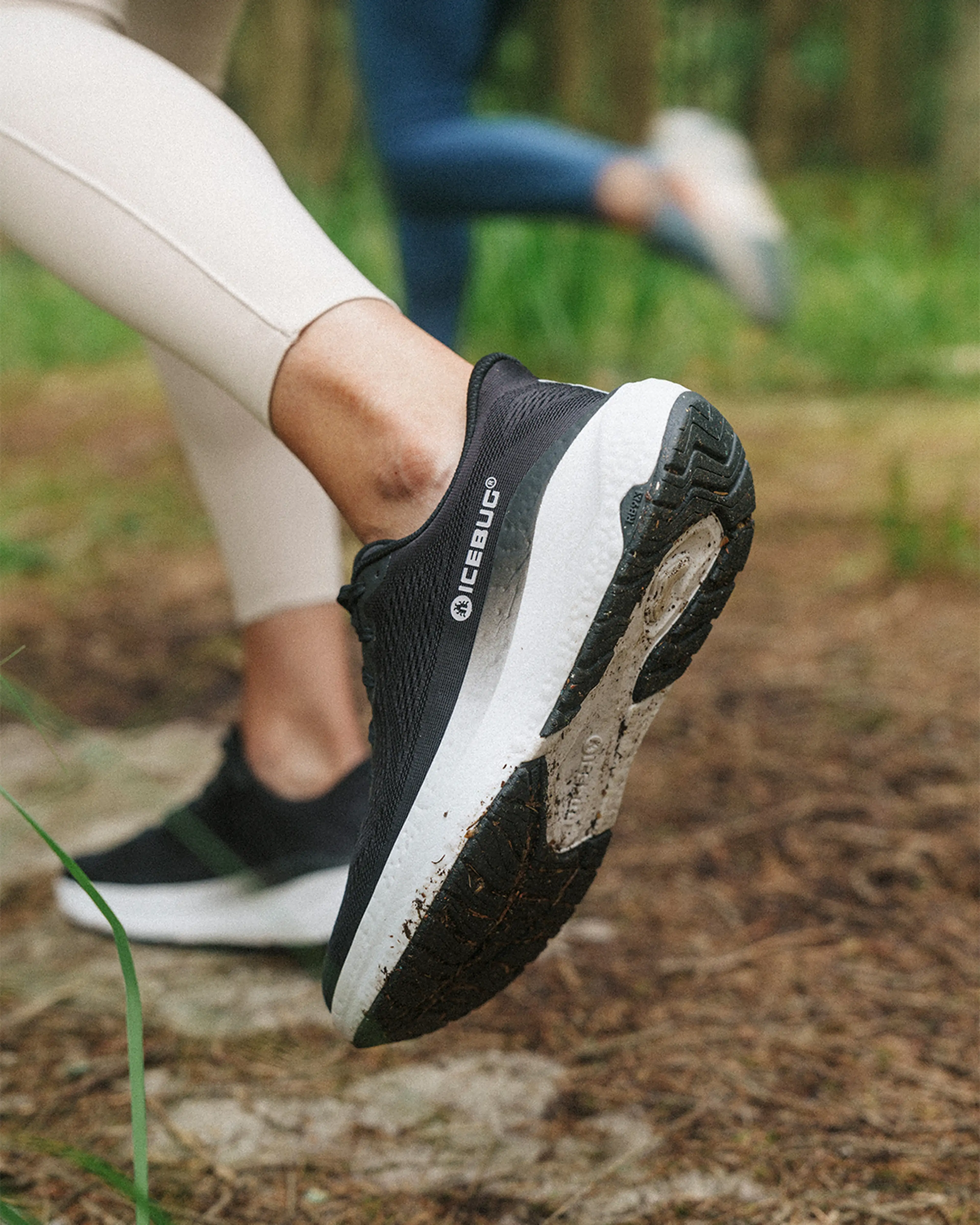 Close-up of a person running on a forest trail, wearing black and white running shoes. Another runner is visible in the background.