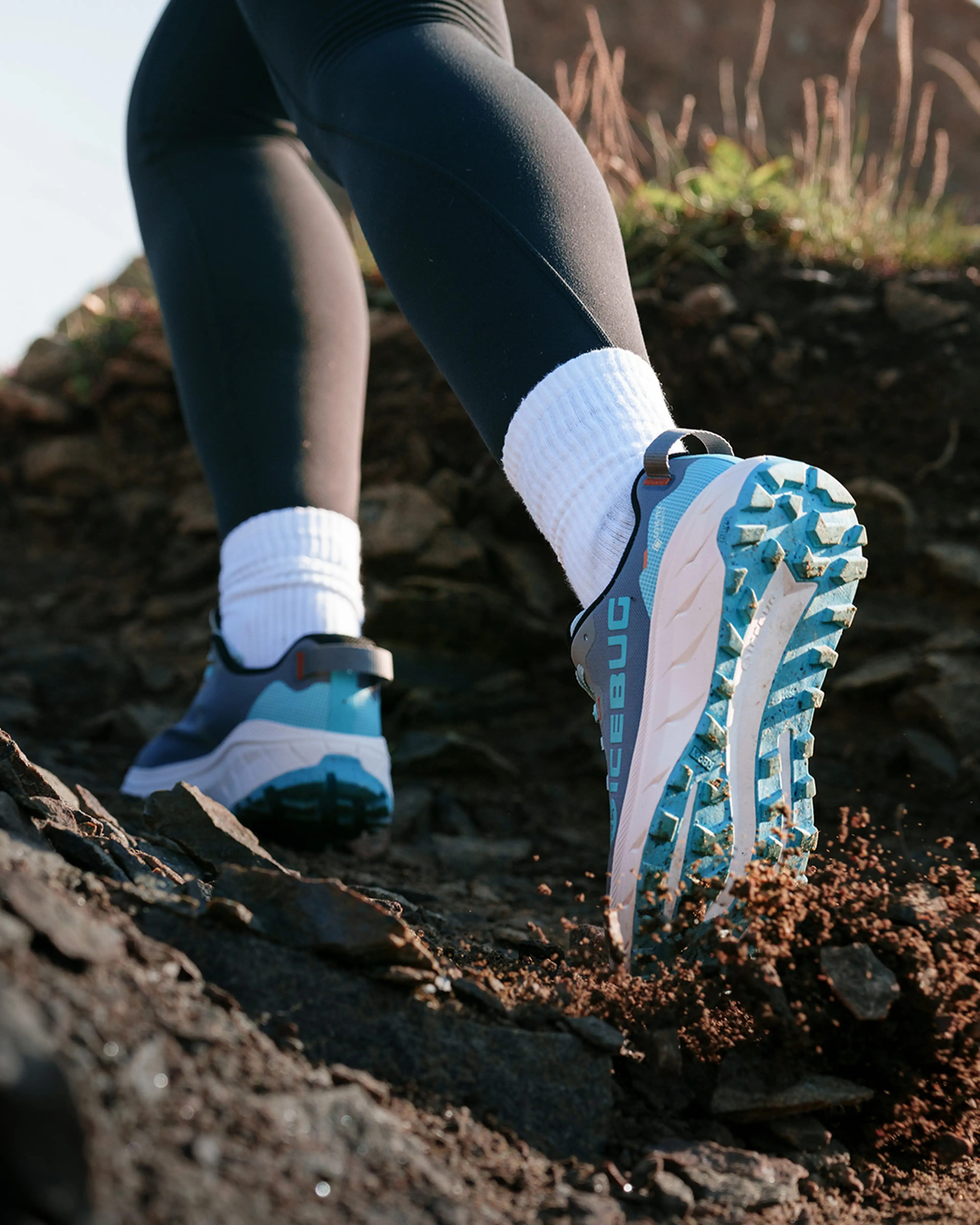 Close-up of a person running on a rocky trail, wearing black leggings, white socks, and blue Icebug trail running shoes with rugged soles.