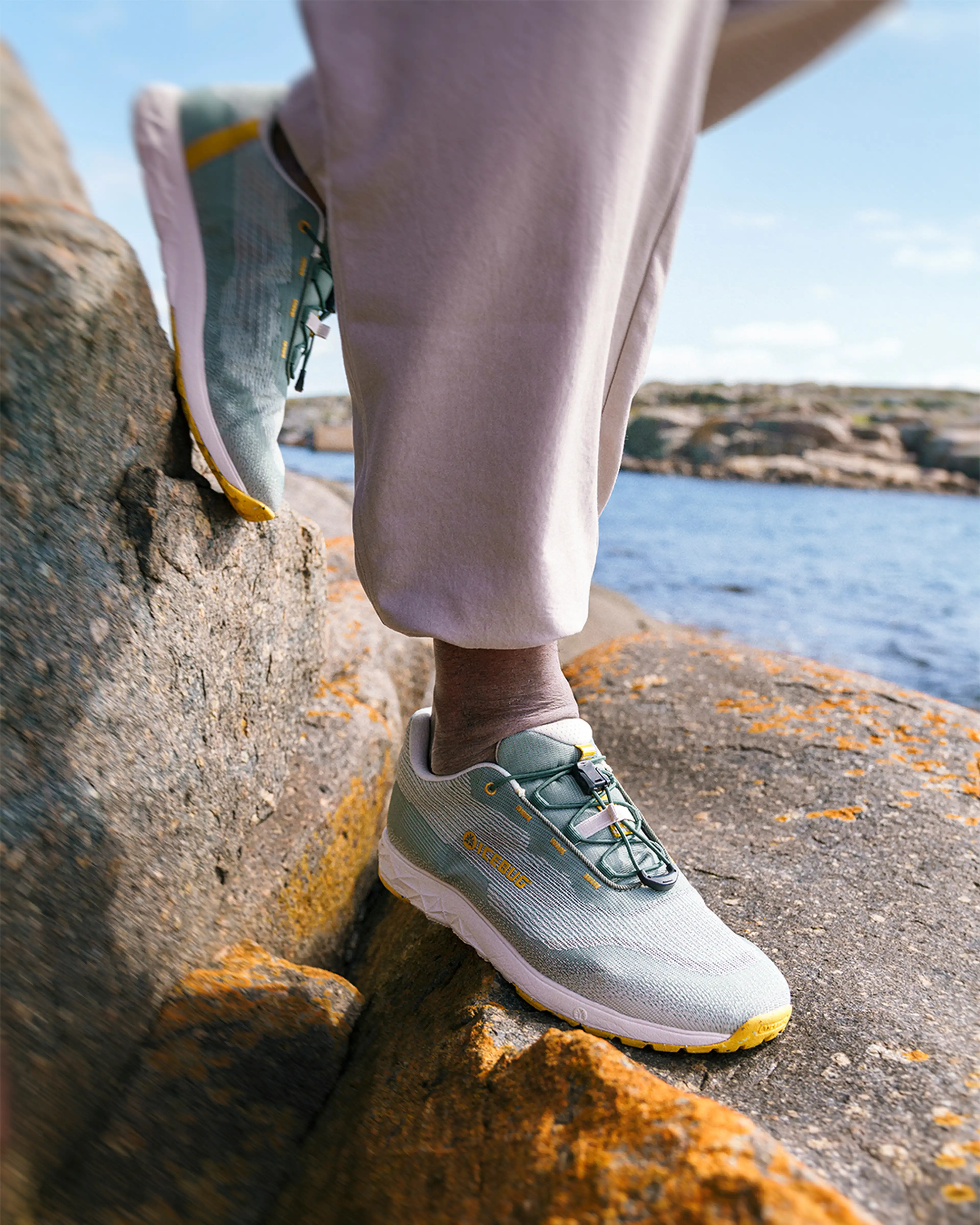 A person wearing light green sneakers and beige pants stands on a rocky surface by the water.