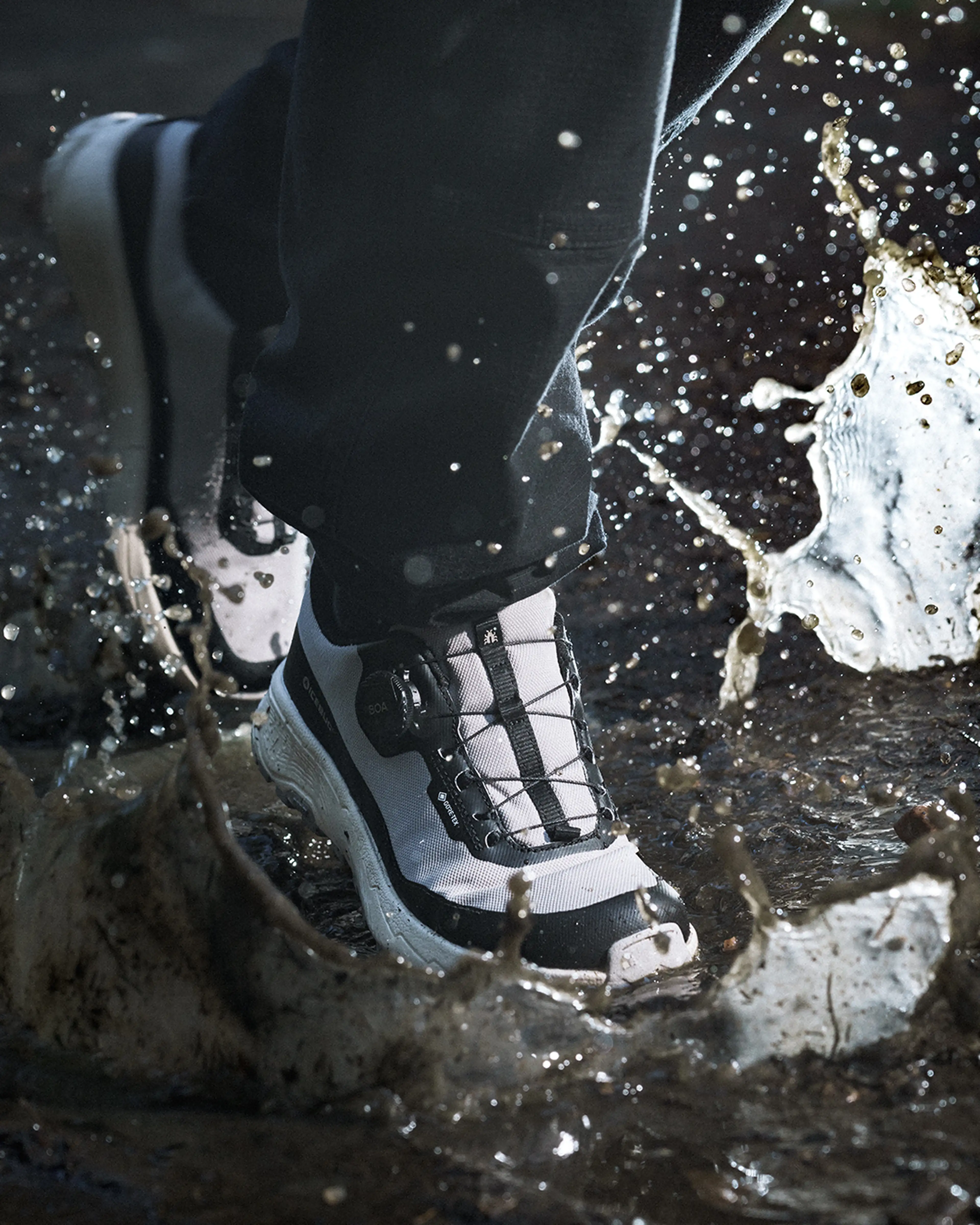 Close-up of a person wearing white and black waterproof shoes splashing through a muddy puddle, causing water to splash around.