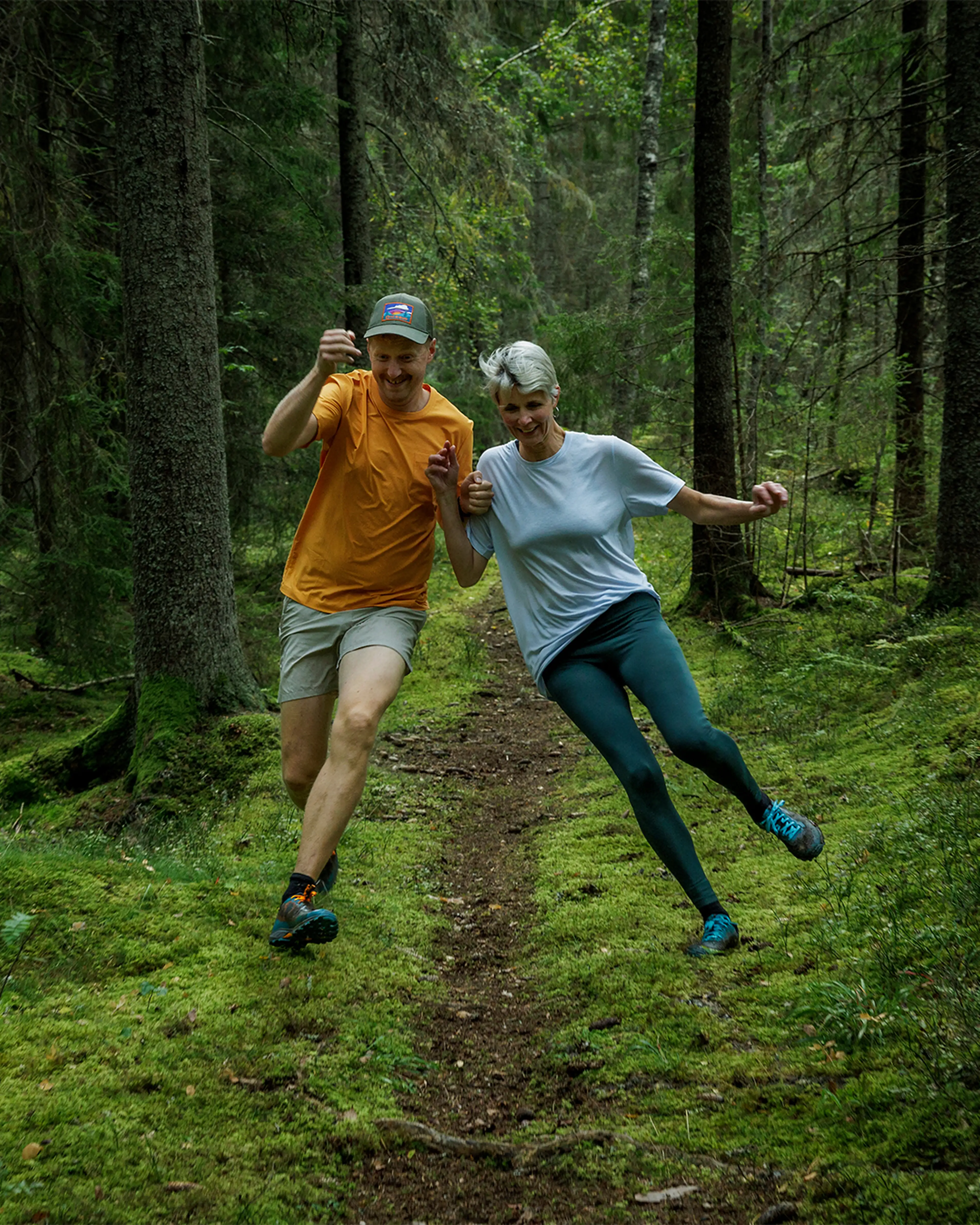 Two people joyfully skipping down a forest path, surrounded by tall trees and lush greenery.