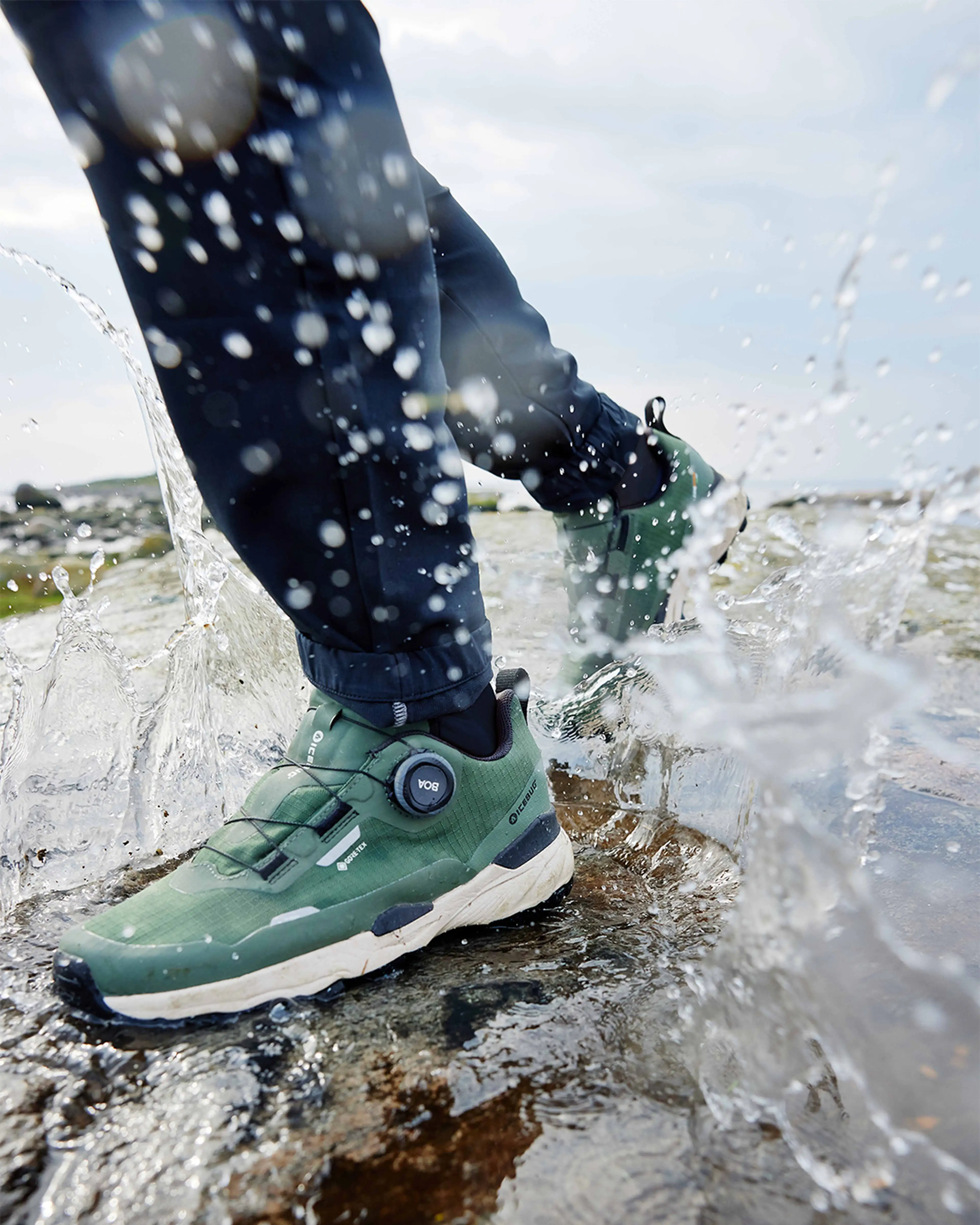 Person wearing green waterproof hiking shoes splashes through a shallow puddle, with water droplets frozen mid-air.