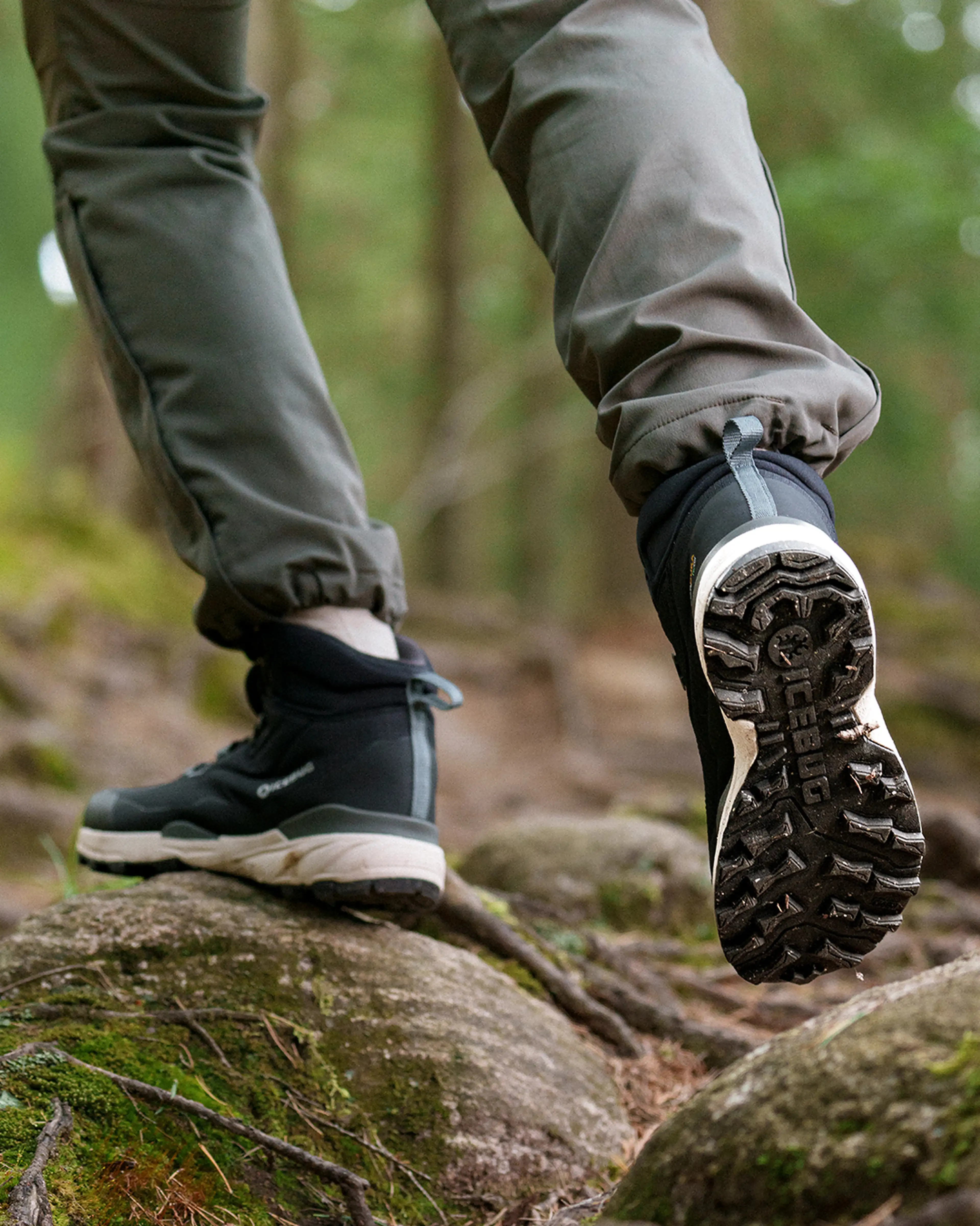 Close-up of a person hiking in the forest, wearing grey pants and black hiking boots with rugged soles, stepping on mossy rocks.