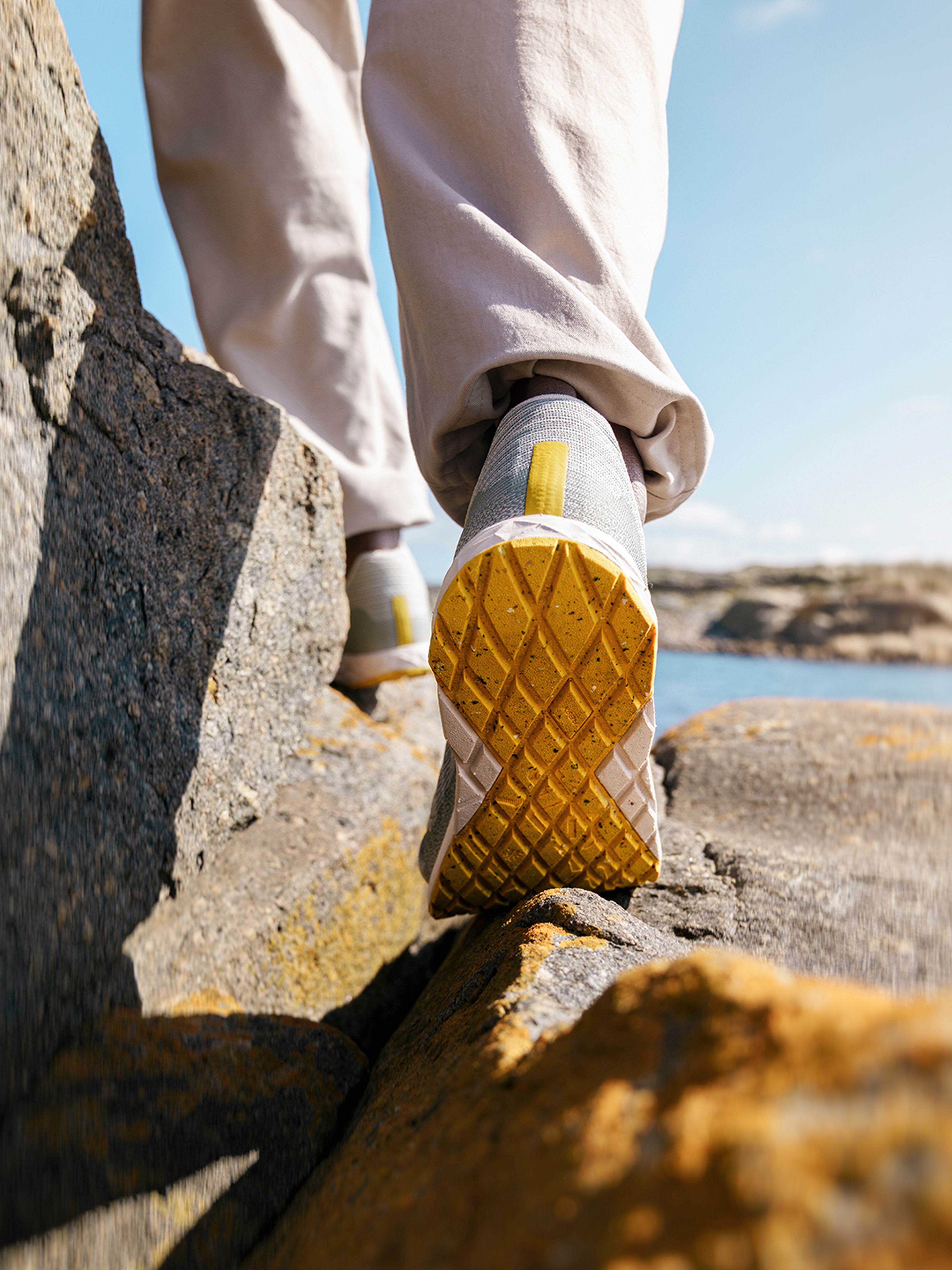 Close-up of a person wearing light green athletic Icebug shoes with yellow soles, walking on rocky terrain by the water under a clear sky.