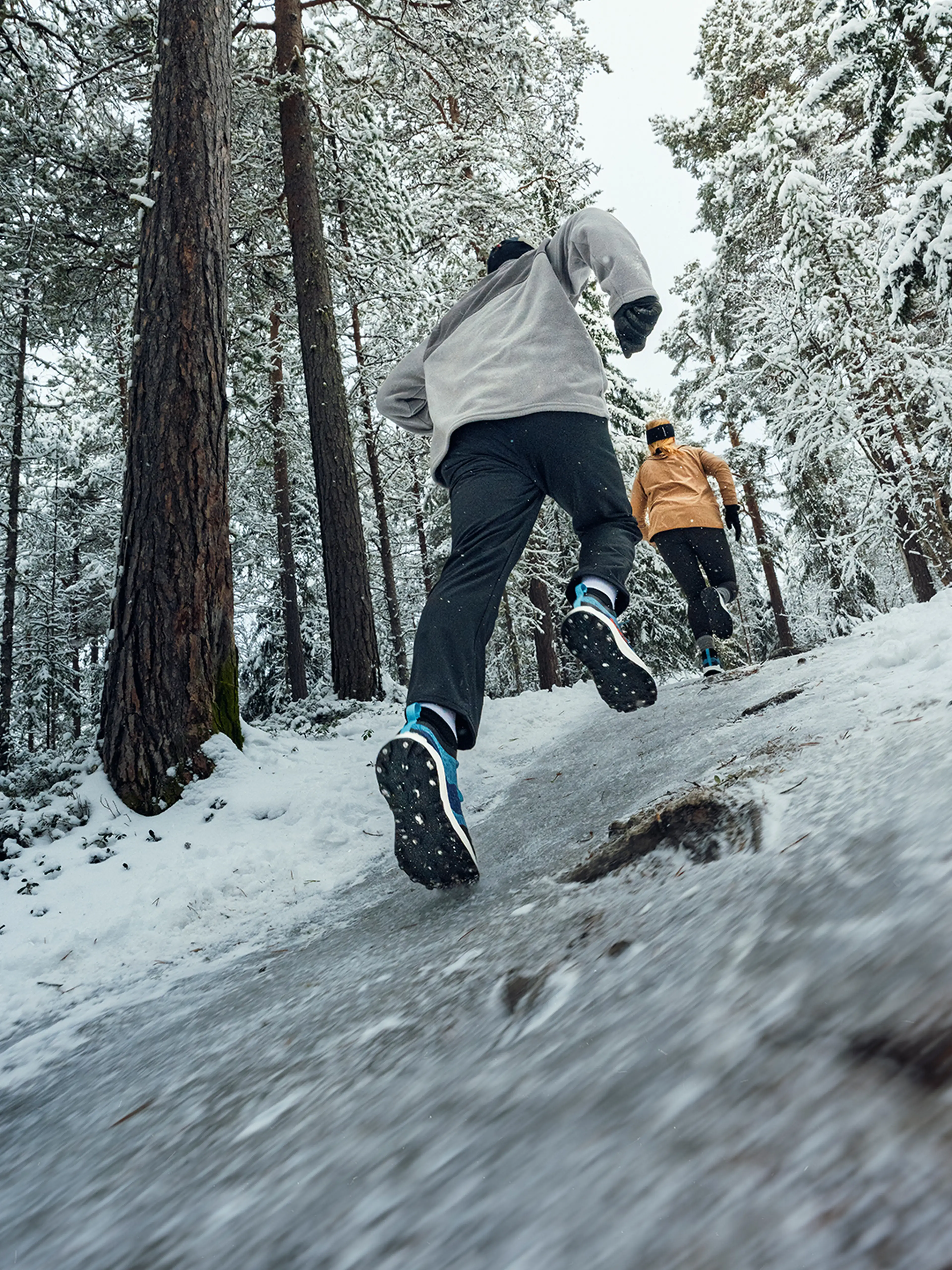 Two people running uphill on a snowy forest trail, wearing winter gear and Icebug studded winter running shoes. Snow-covered trees surround them.