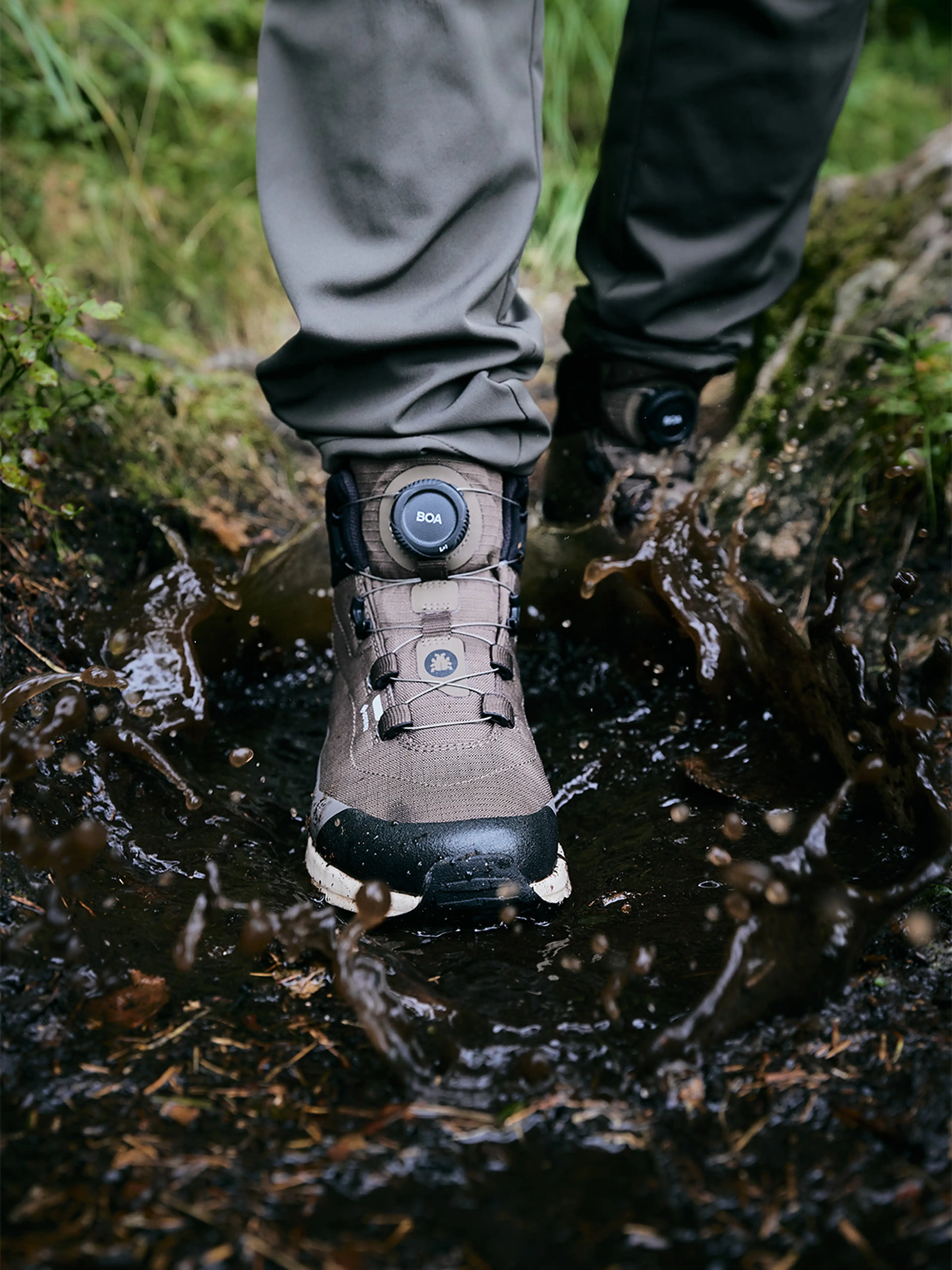A close-up of a person wearing waterproof hiking boots stepping in a muddy puddle, causing splashes, with green foliage in the background.