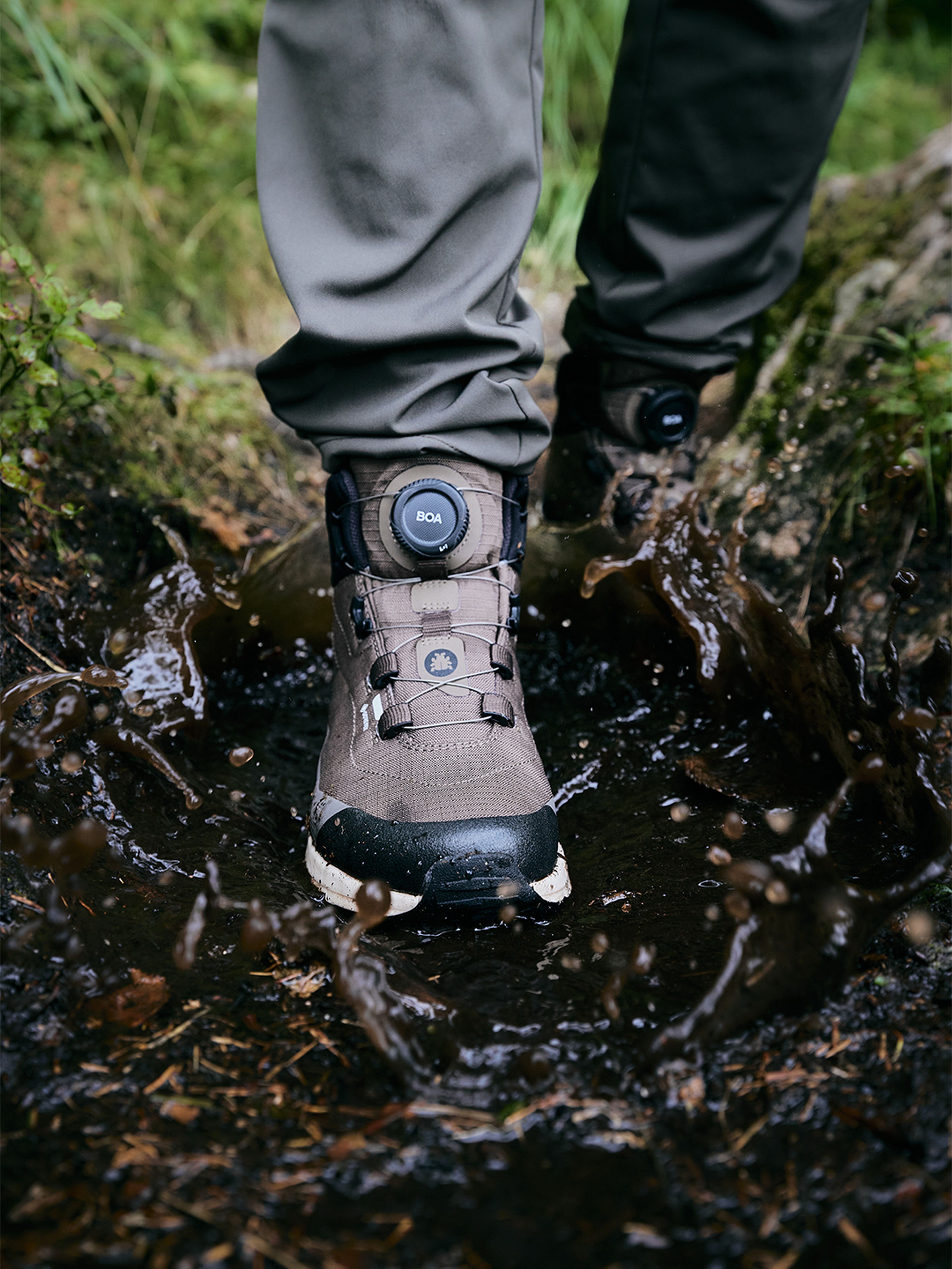 A person wearing waterproof hiking boots steps into a muddy puddle, causing water to splash around.