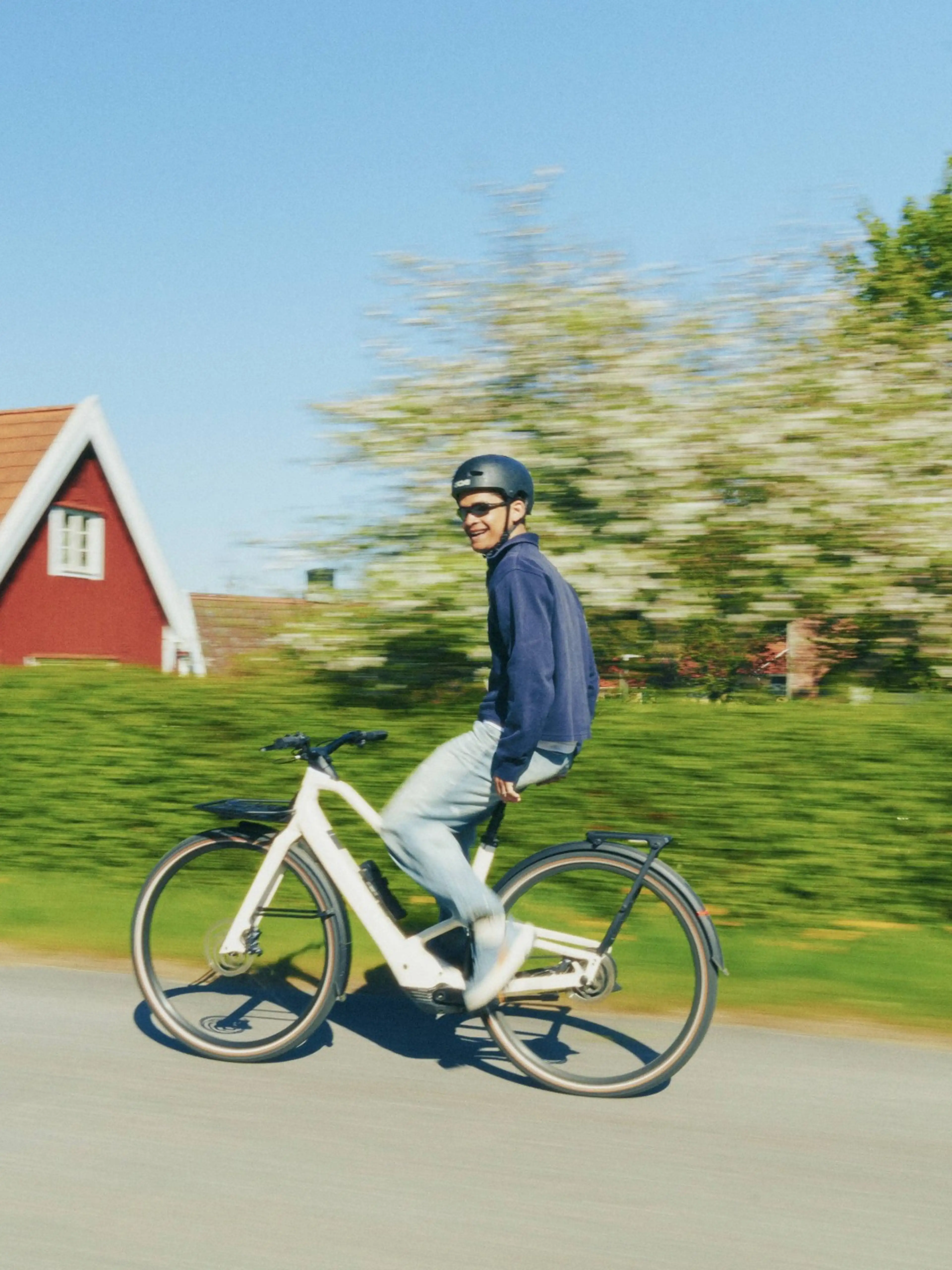 A person wearing a helmet rides a white bicycle on a sunny day, passing a red house and green hedges, with motion blur in the background.