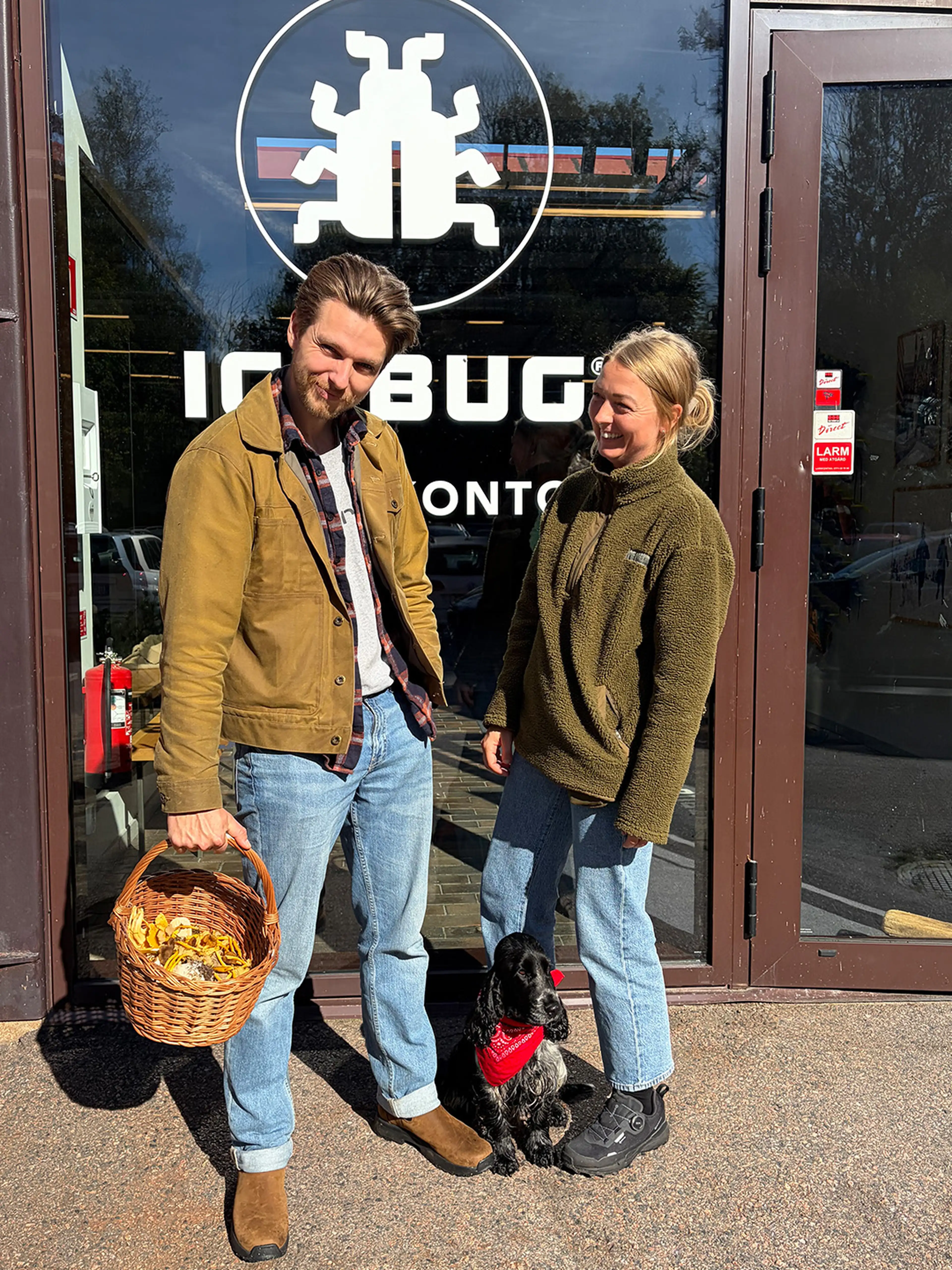 A man and woman stand outside Icebug HQ, holding a basket. A dog with a red bandana sits between them.