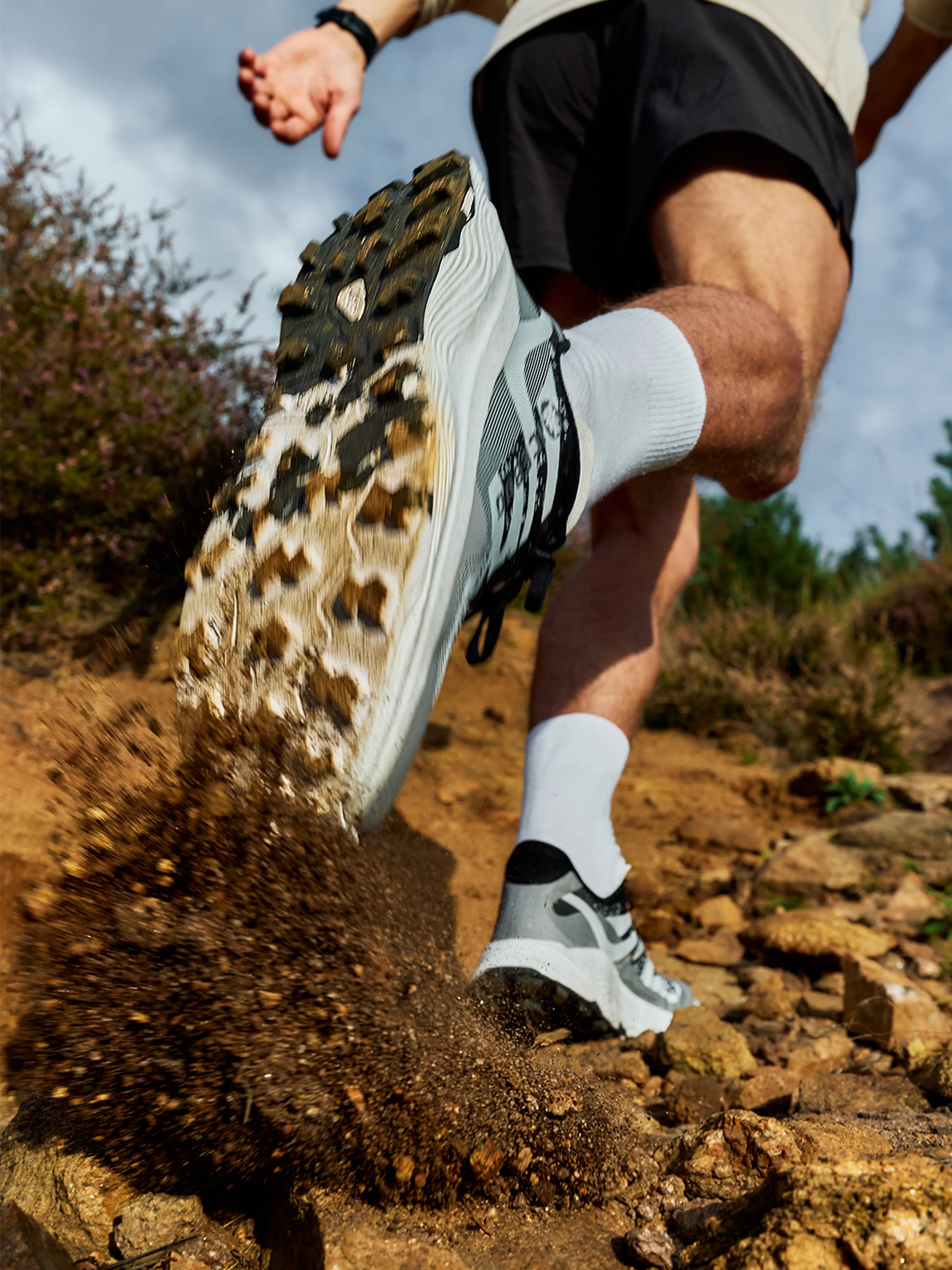 Close-up of a person running uphill on a rocky trail, kicking up dirt in white socks and athletic shoes, with a cloudy sky in the background.