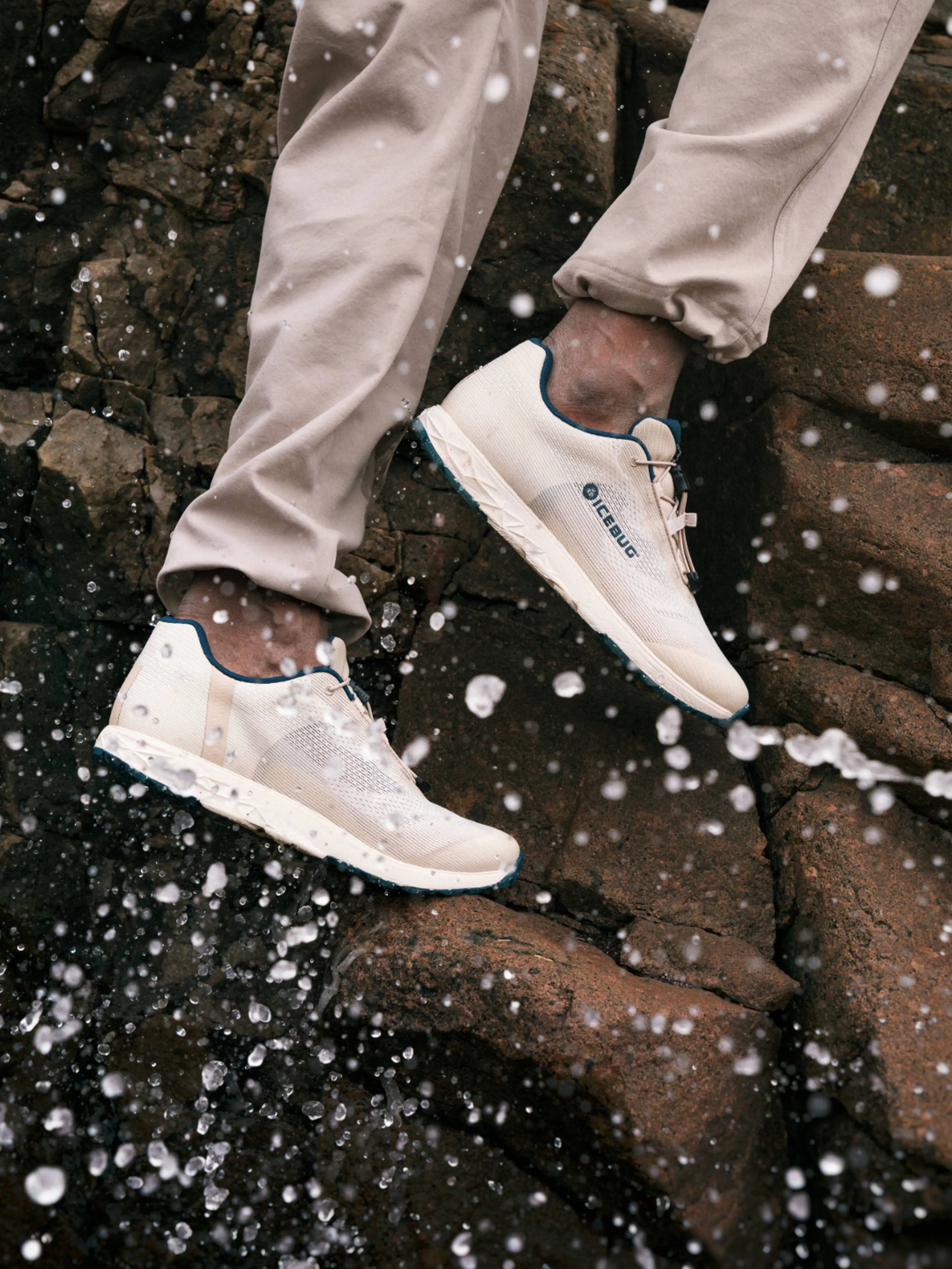 Person wearing beige pants and light sneakers on wet rocks, with splashing water droplets around.