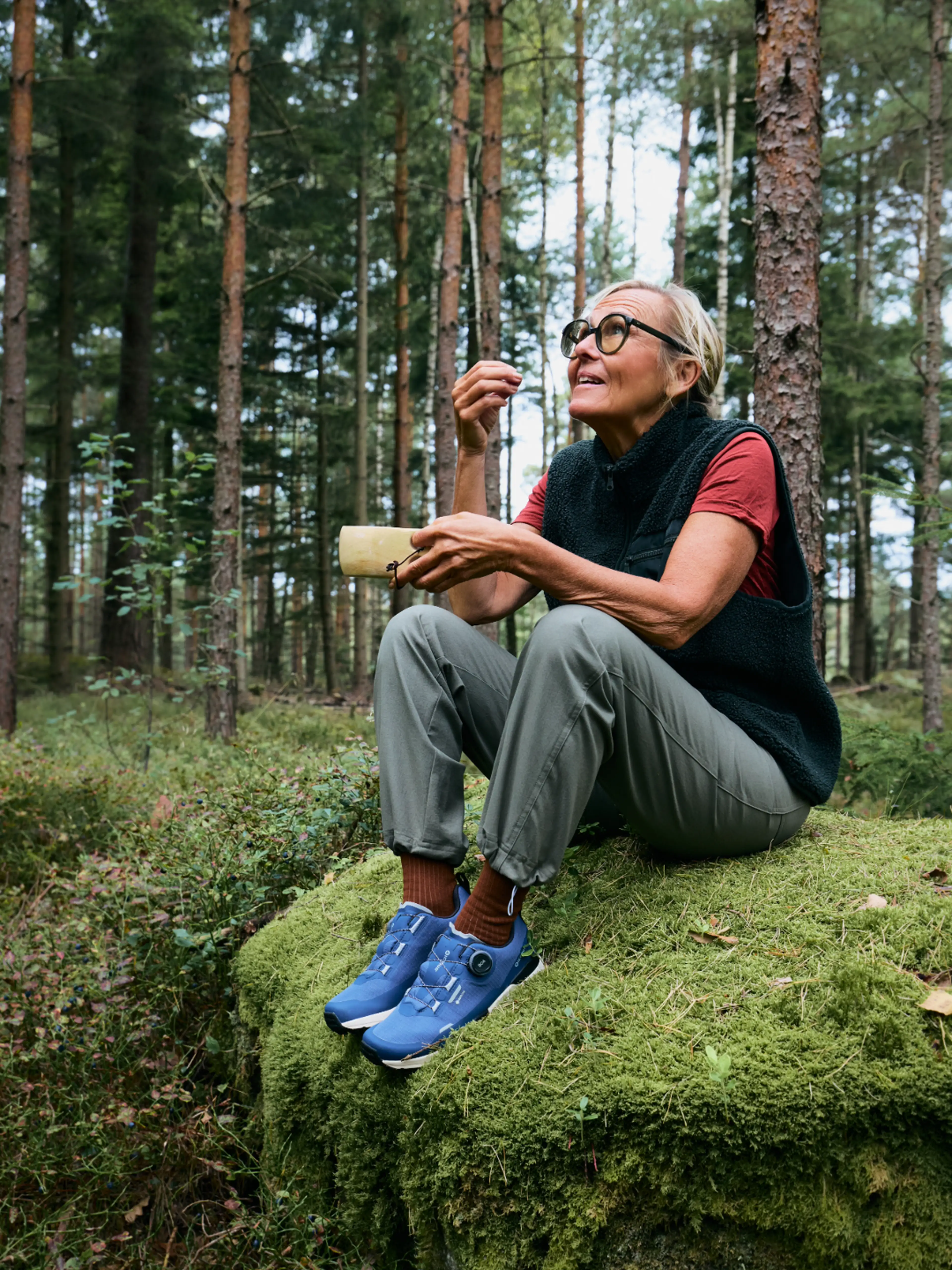 Woman sitting on a mossy rock in a forest, sipping from a cup and smiling, surrounded by tall trees.