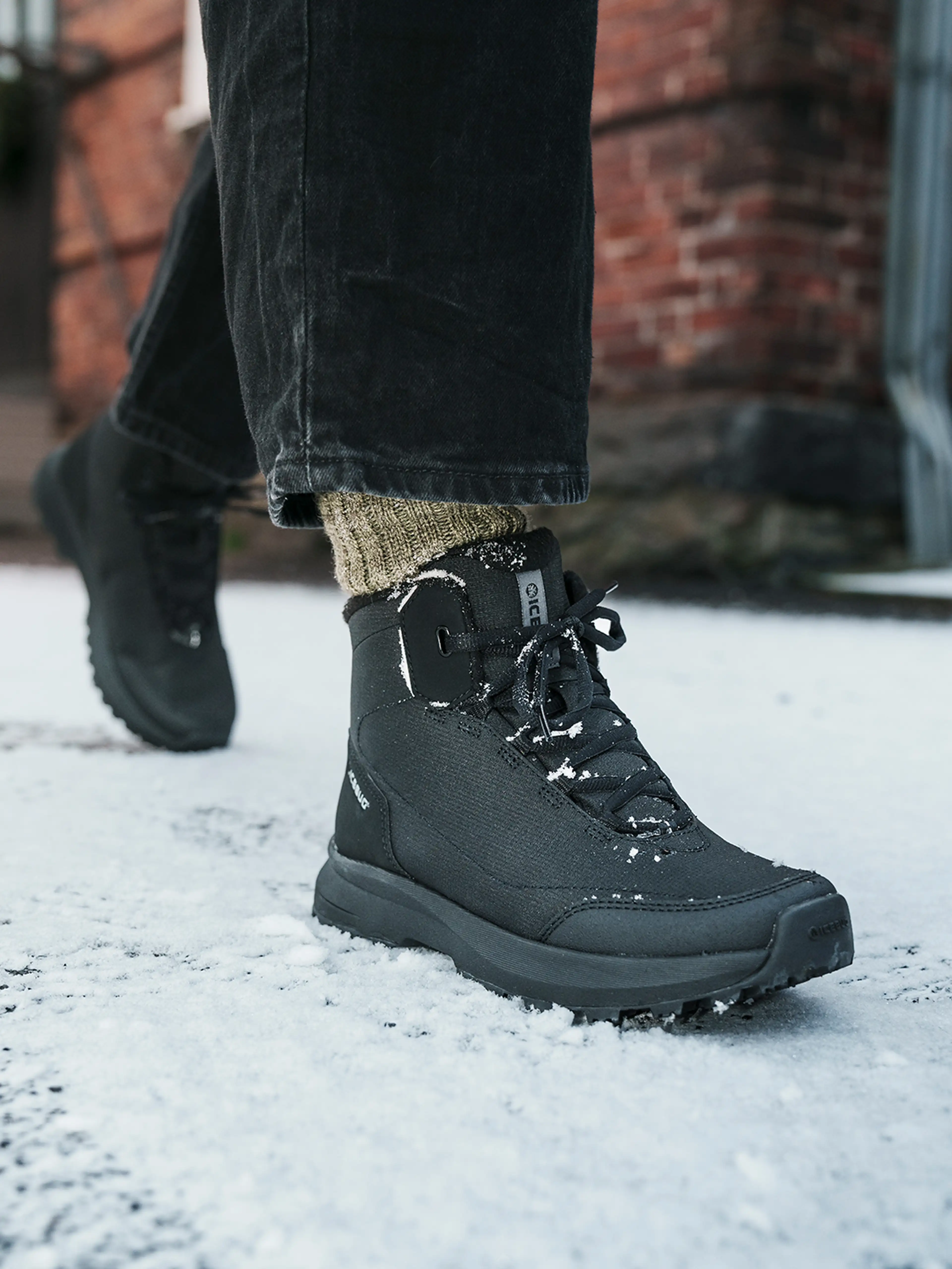 Person wearing black boots and dark pants walking on snow-covered ground, with a brick building in the background.