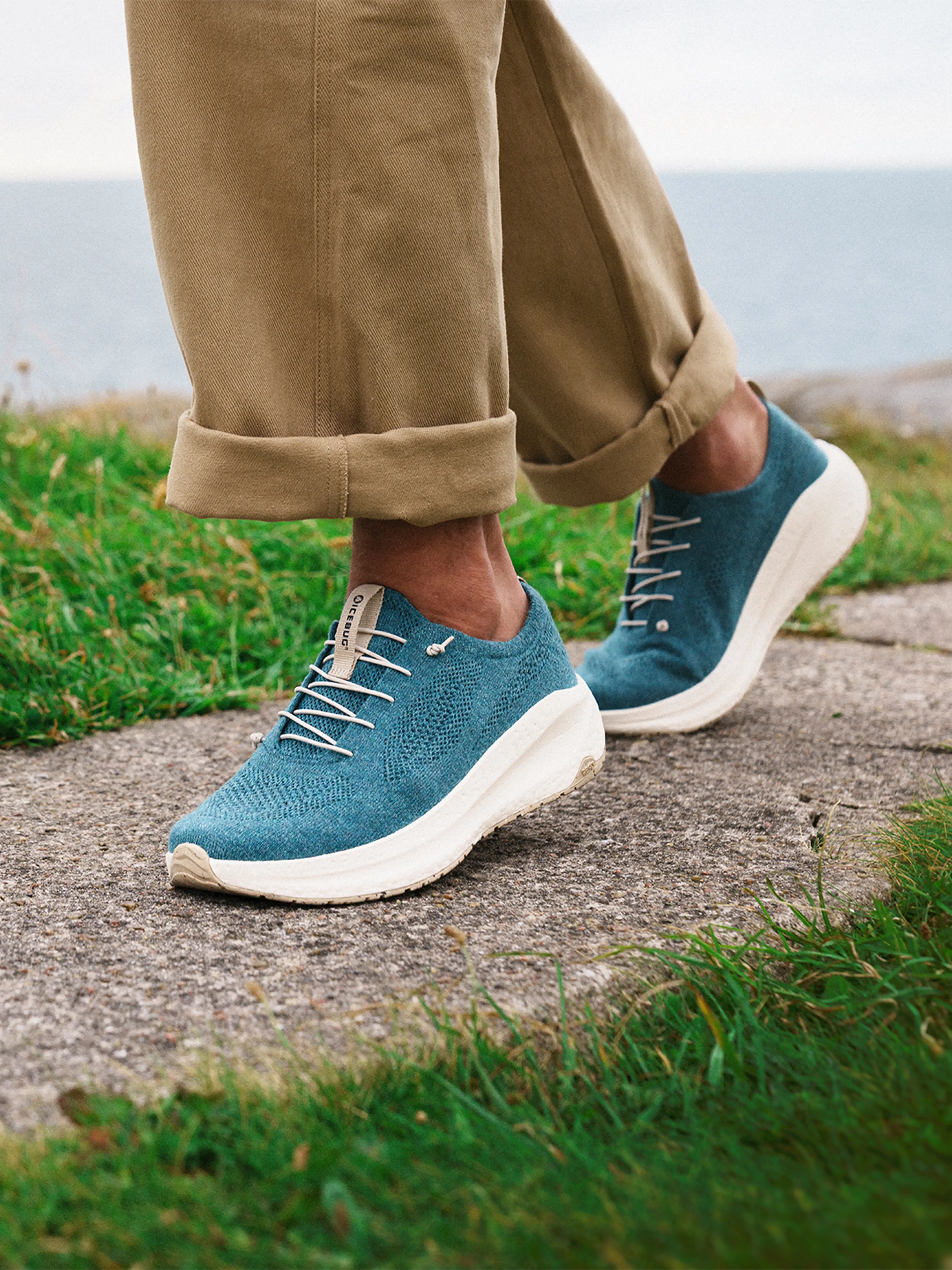Person wearing blue Icebug sneakers with white laces, beige pants rolled up, standing on a stone path with grass and ocean in the background.