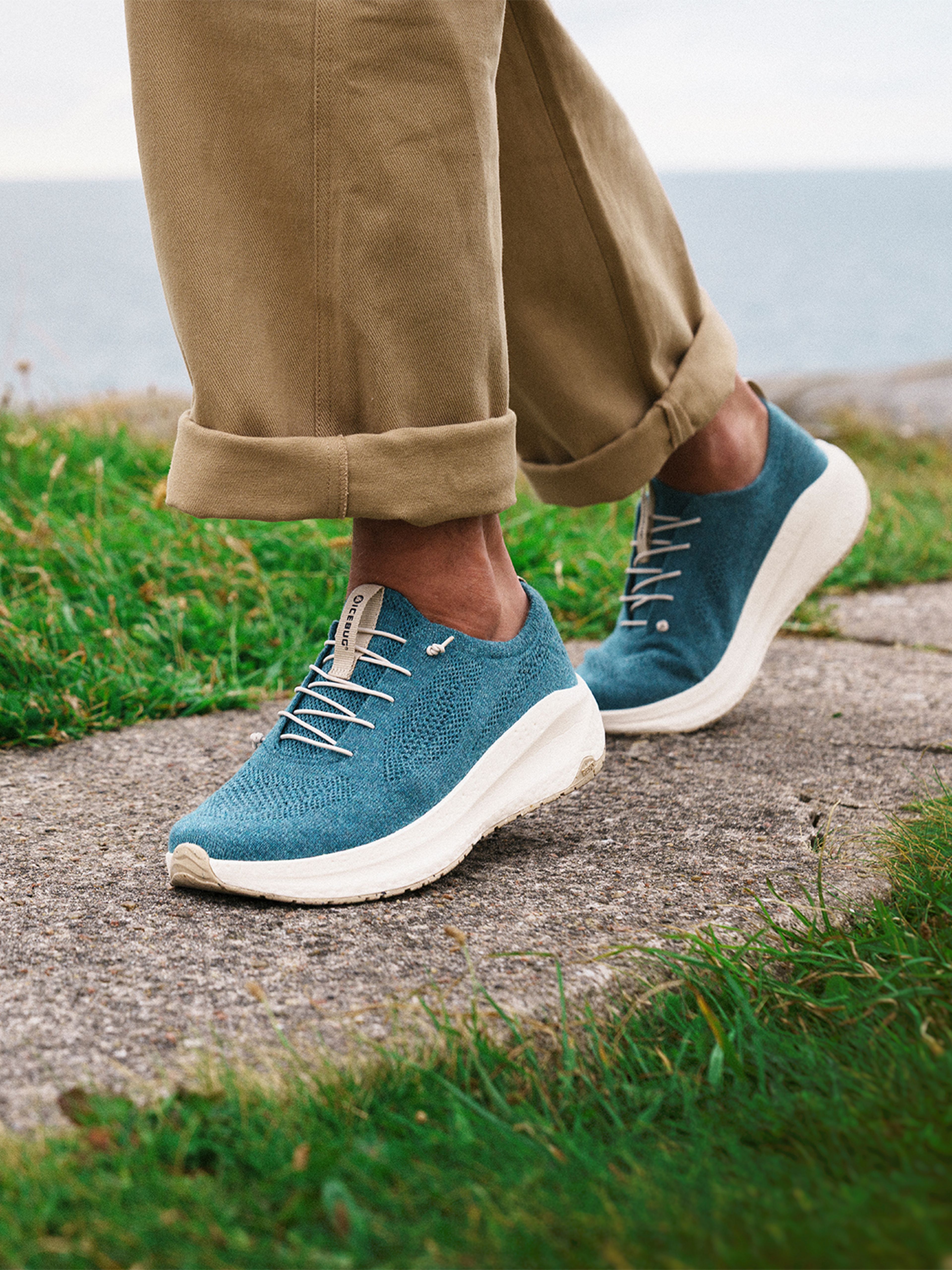 Person wearing blue Icebug sneakers with white laces, beige pants rolled up, standing on a stone path with grass and ocean in the background.
