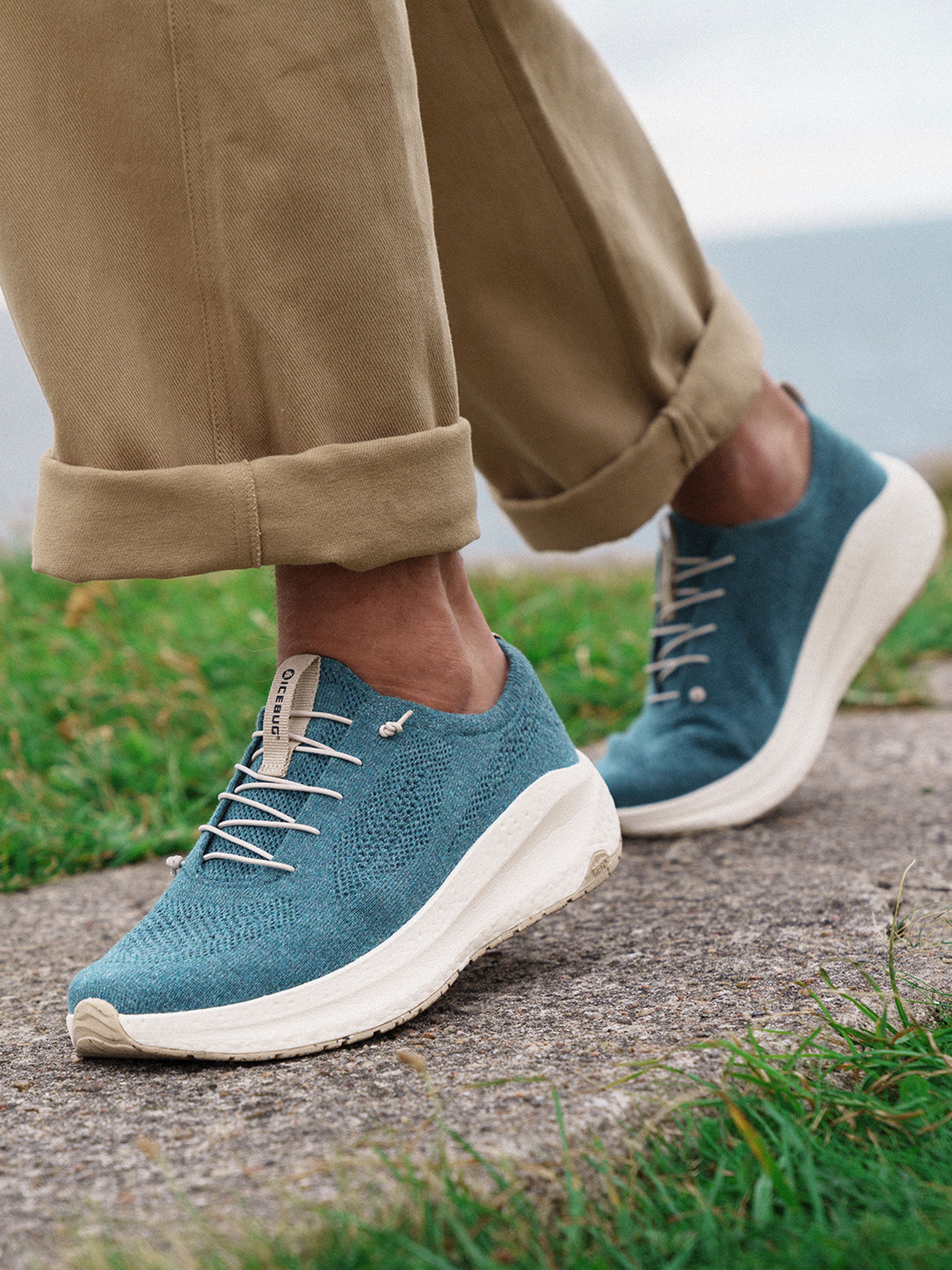 Person wearing blue Icebug sneakers with white laces, beige pants rolled up, standing on a stone path with grass and ocean in the background.