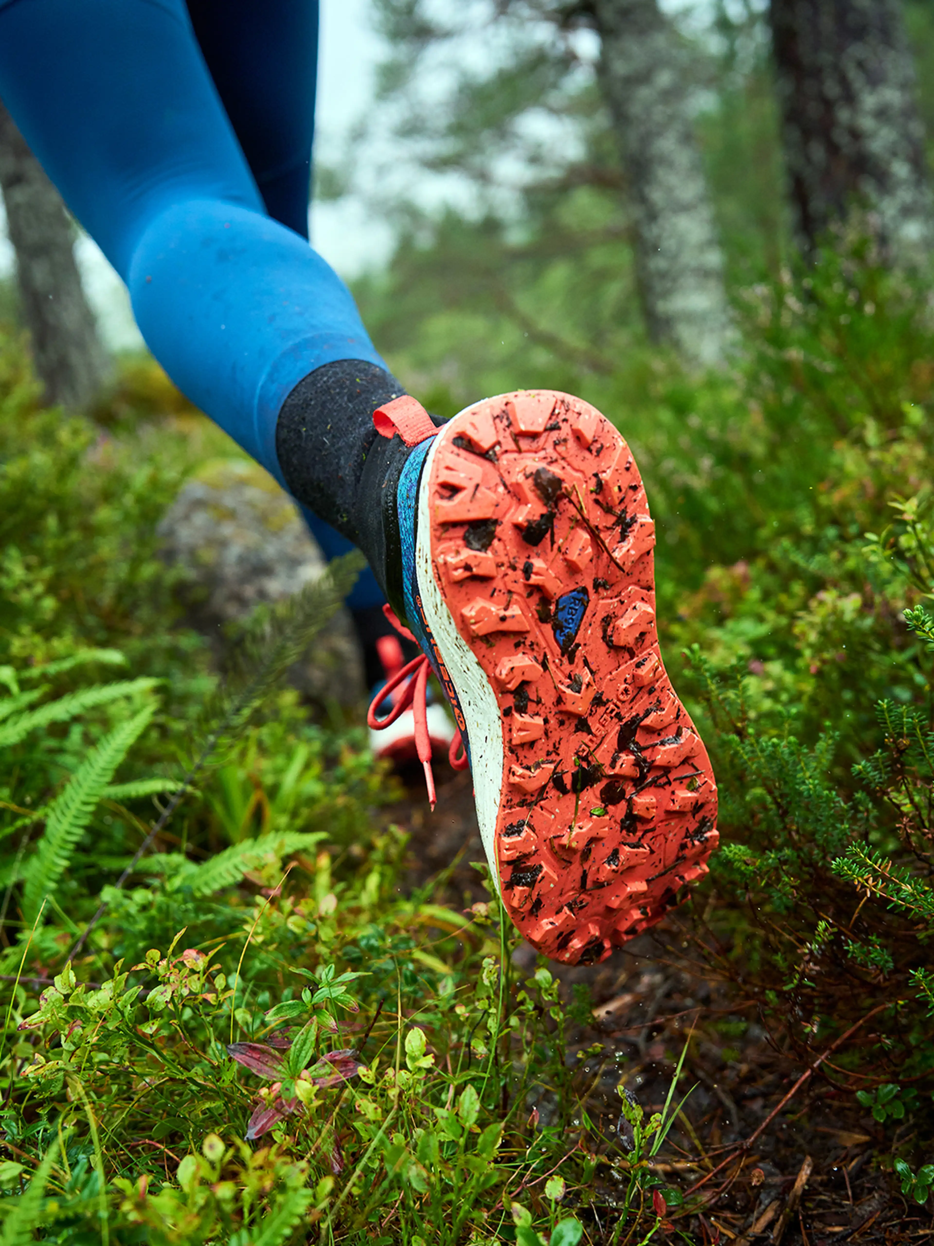 Close-up of a person wearing blue leggings and Icebug trail running shoes with orange soles, running on a muddy forest trail surrounded by greenery.