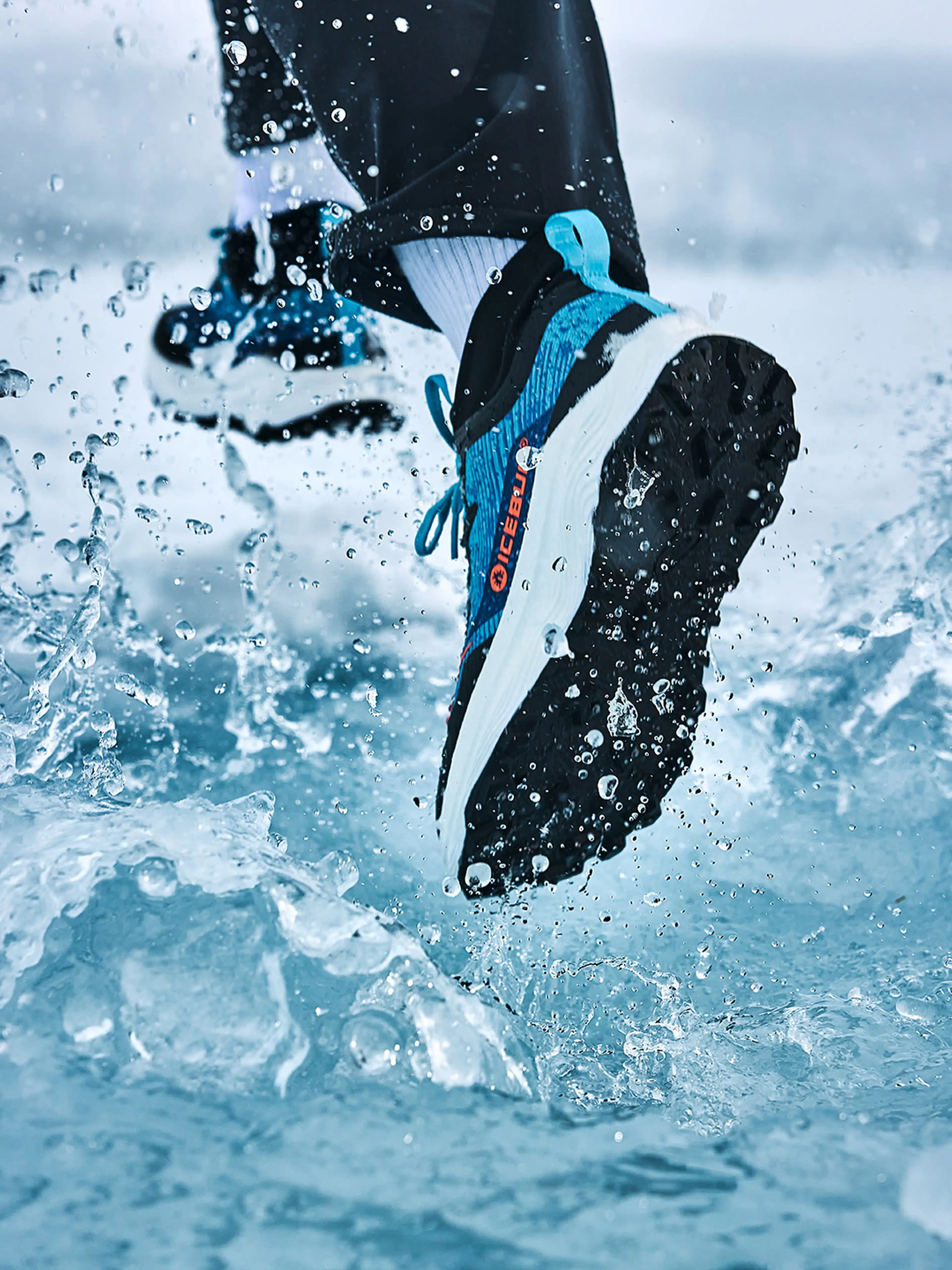 Close-up of a person running through water, wearing black and blue winter running shoes from Icebug, with water splashing around.