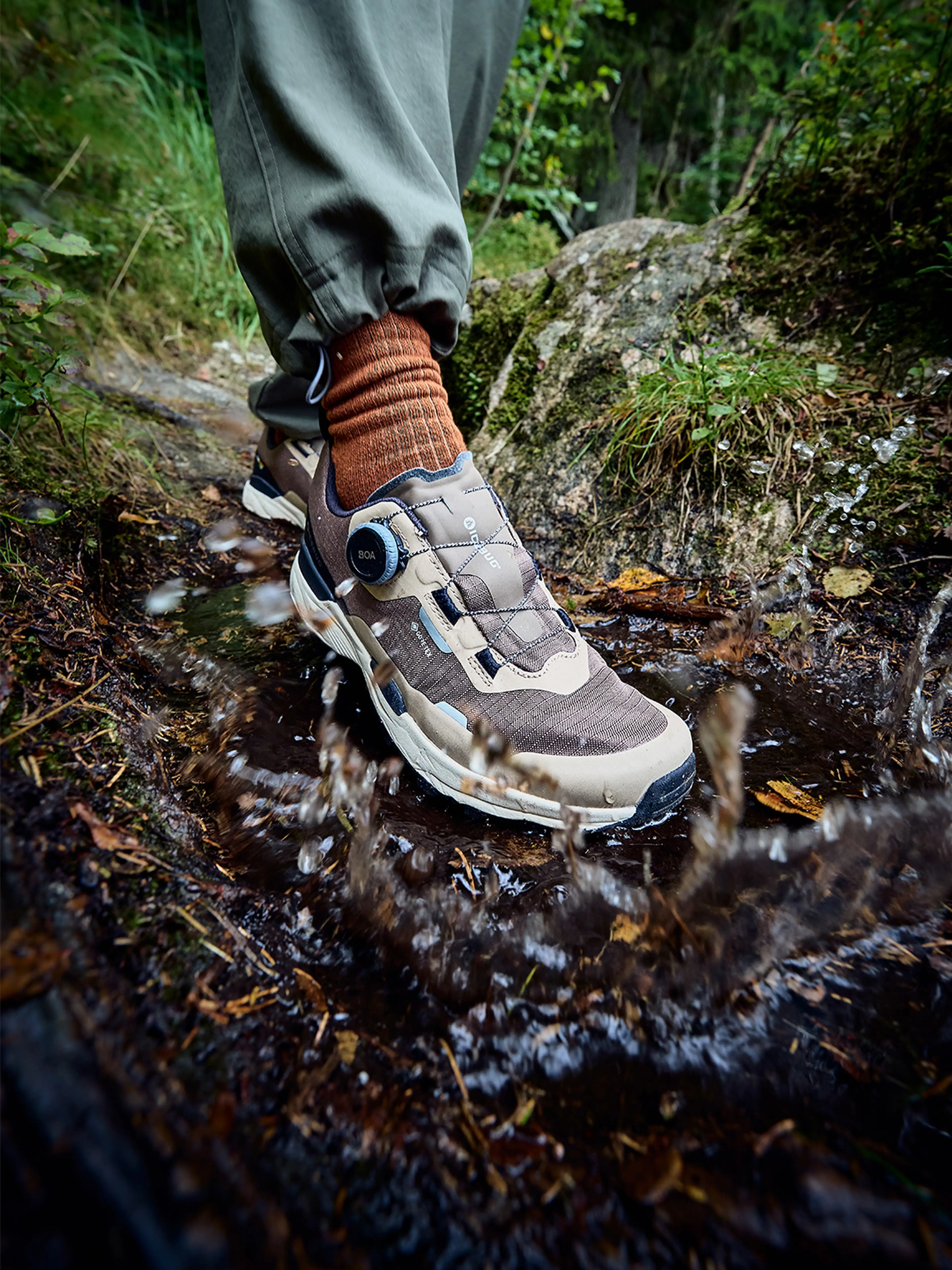 A hiker wearing brown waterproof Icebug hiking shoes and orange socks steps through a muddy puddle on a forest trail, splashing water.