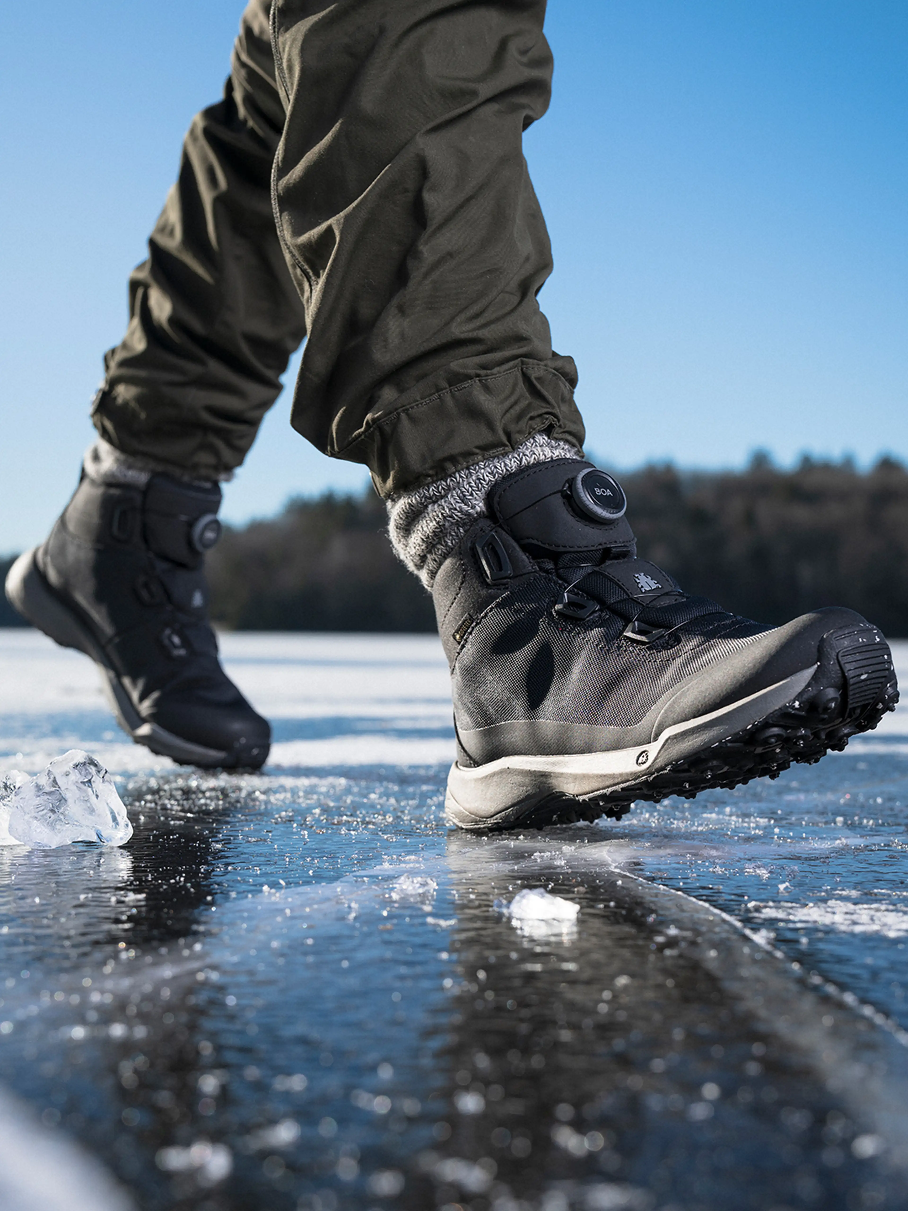 Close-up of a person walking on ice wearing black winter boots, gray socks, and olive green pants, with a clear blue sky background.