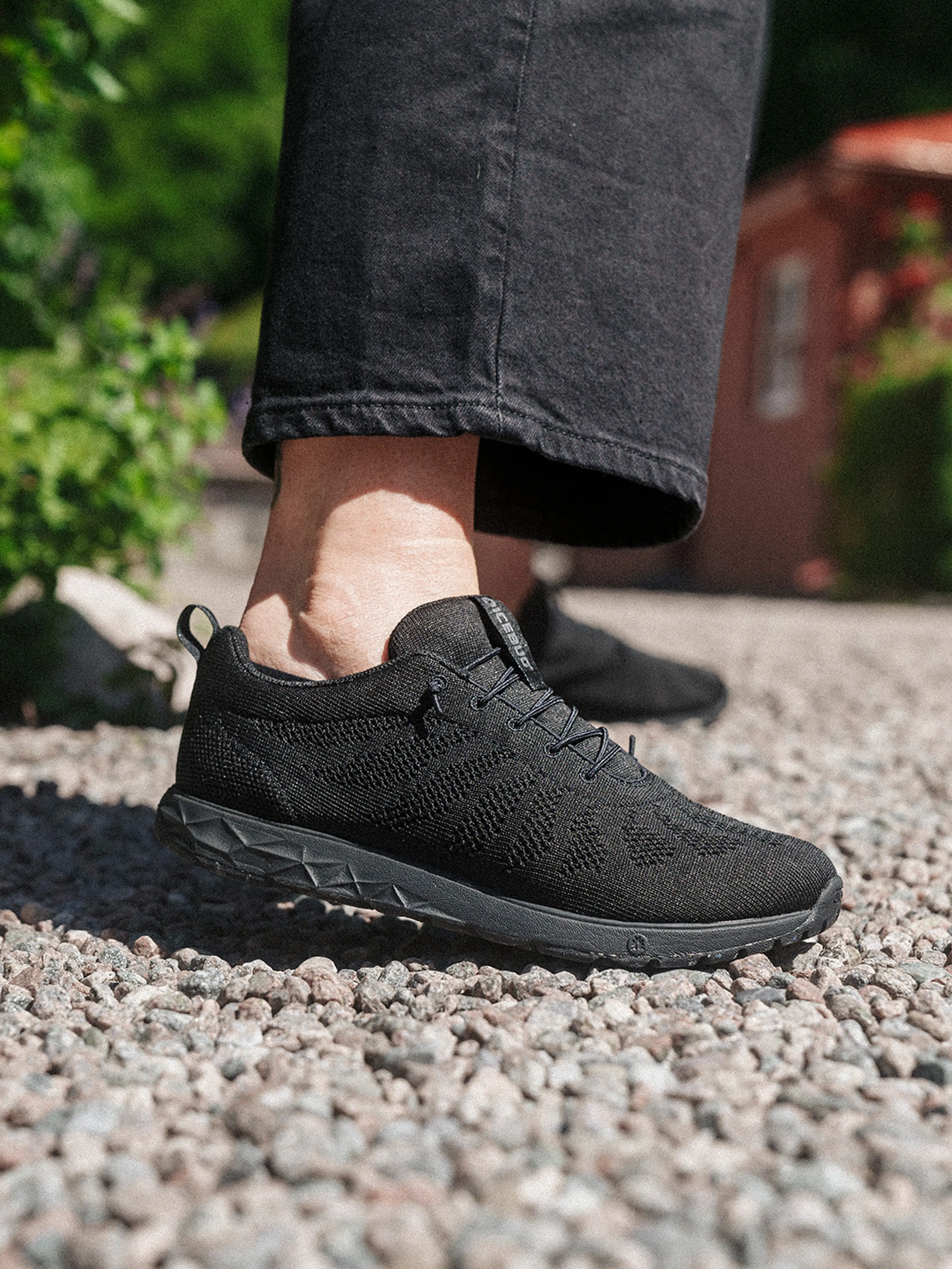 Person wearing black knit sneakers and black pants, standing on gravel outdoors with greenery in the background.