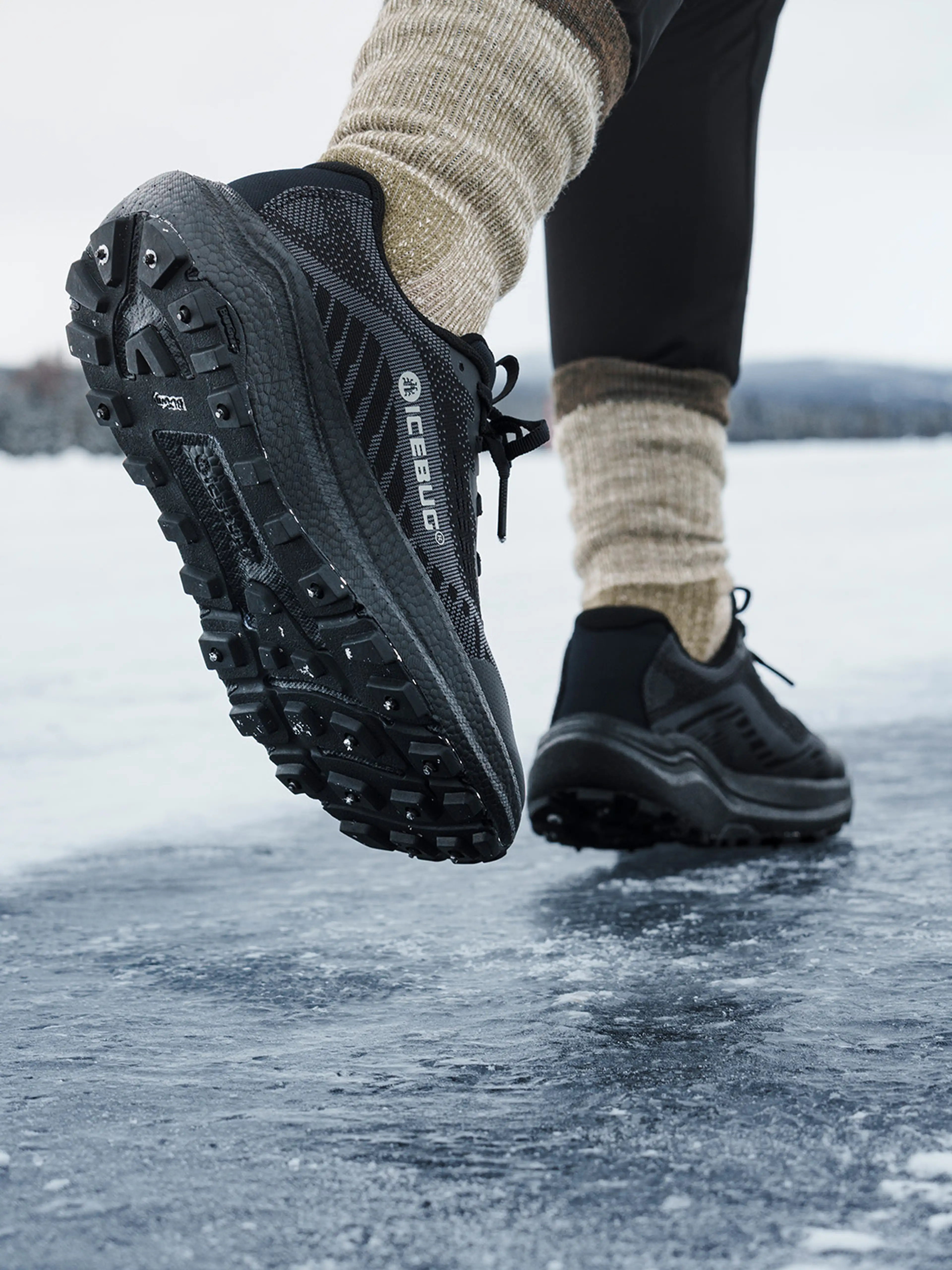 Close-up of a person wearing black trail shoes and beige socks stepping on icy terrain, showcasing the shoe's tread and grip.