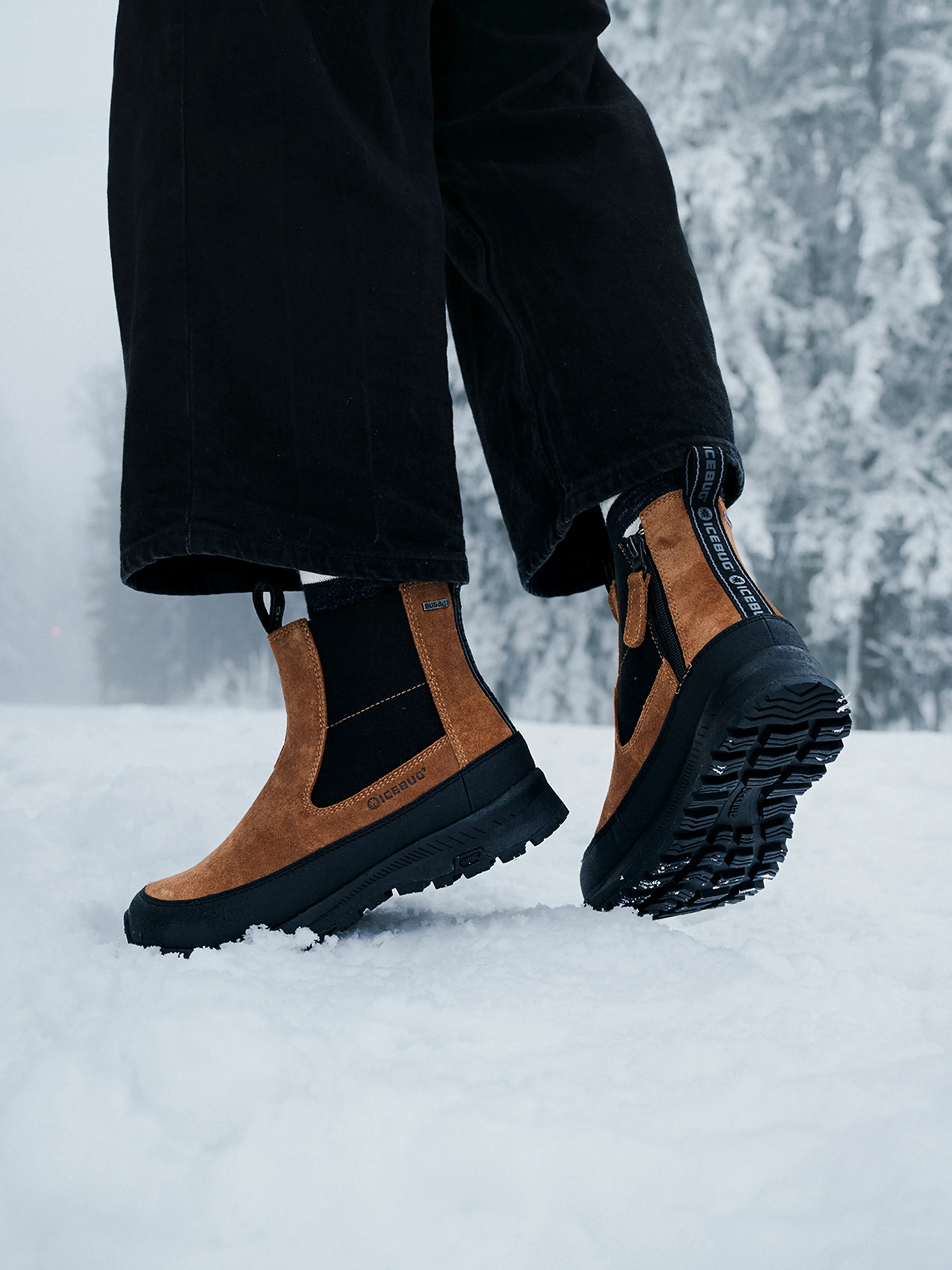 Person wearing brown and black winter boots from Icebug stands on snow, with snowy trees in the background.