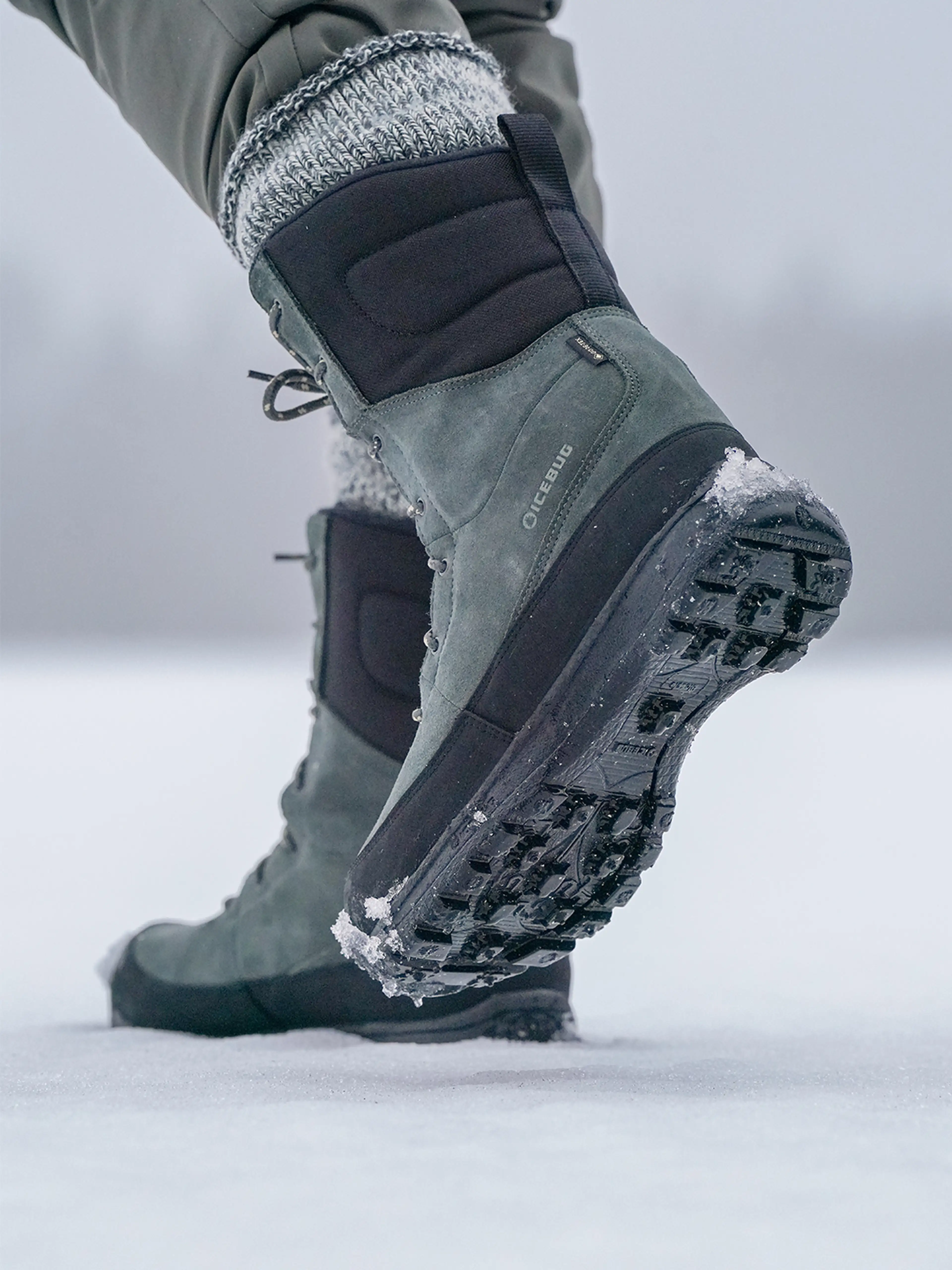 Close-up of a person walking on snow, wearing green and black winter boots with visible tread and snowflakes on the soles.