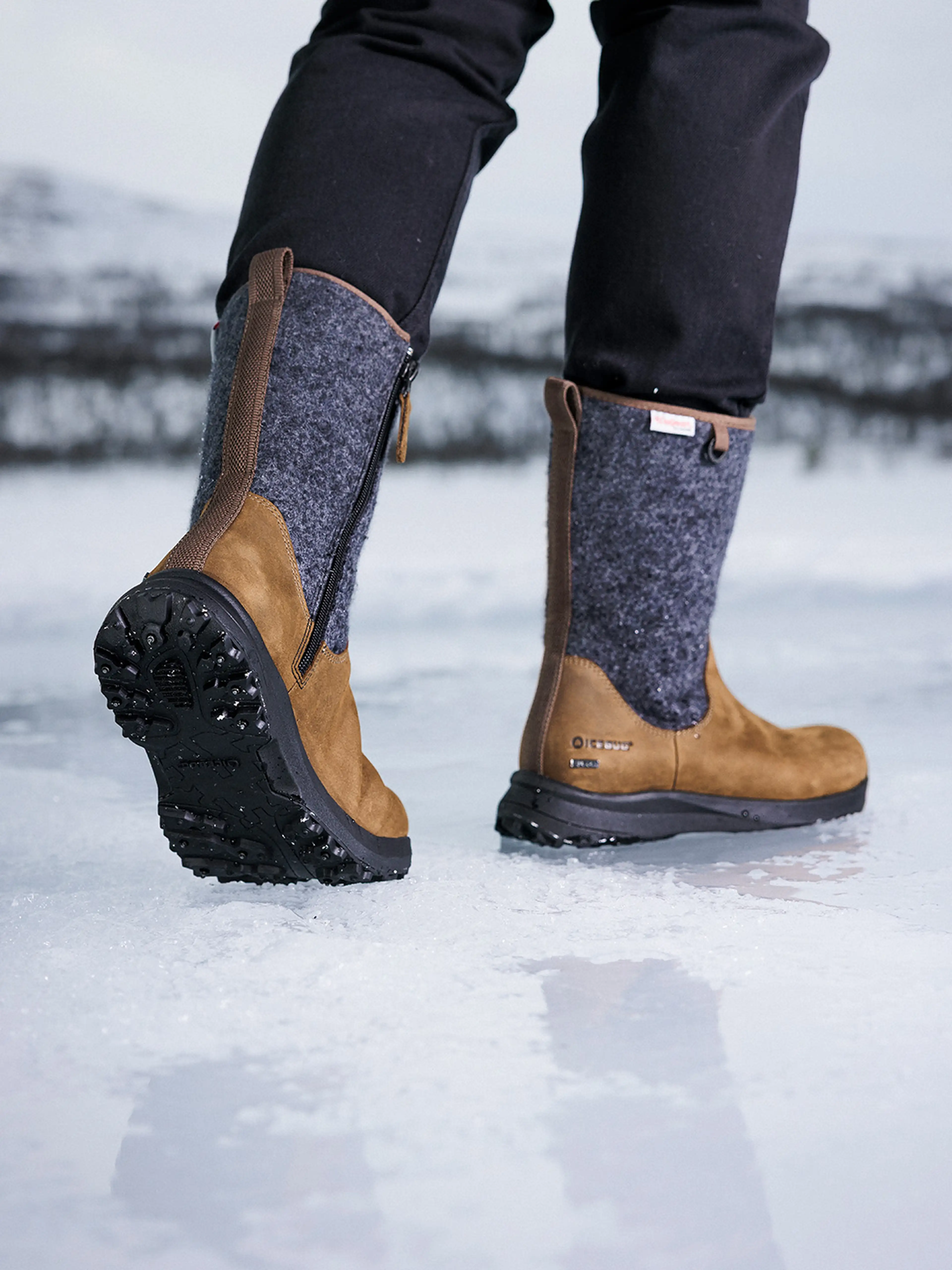 Close-up of a person wearing brown and gray winter boots walking on ice, with snowy landscape in the background.