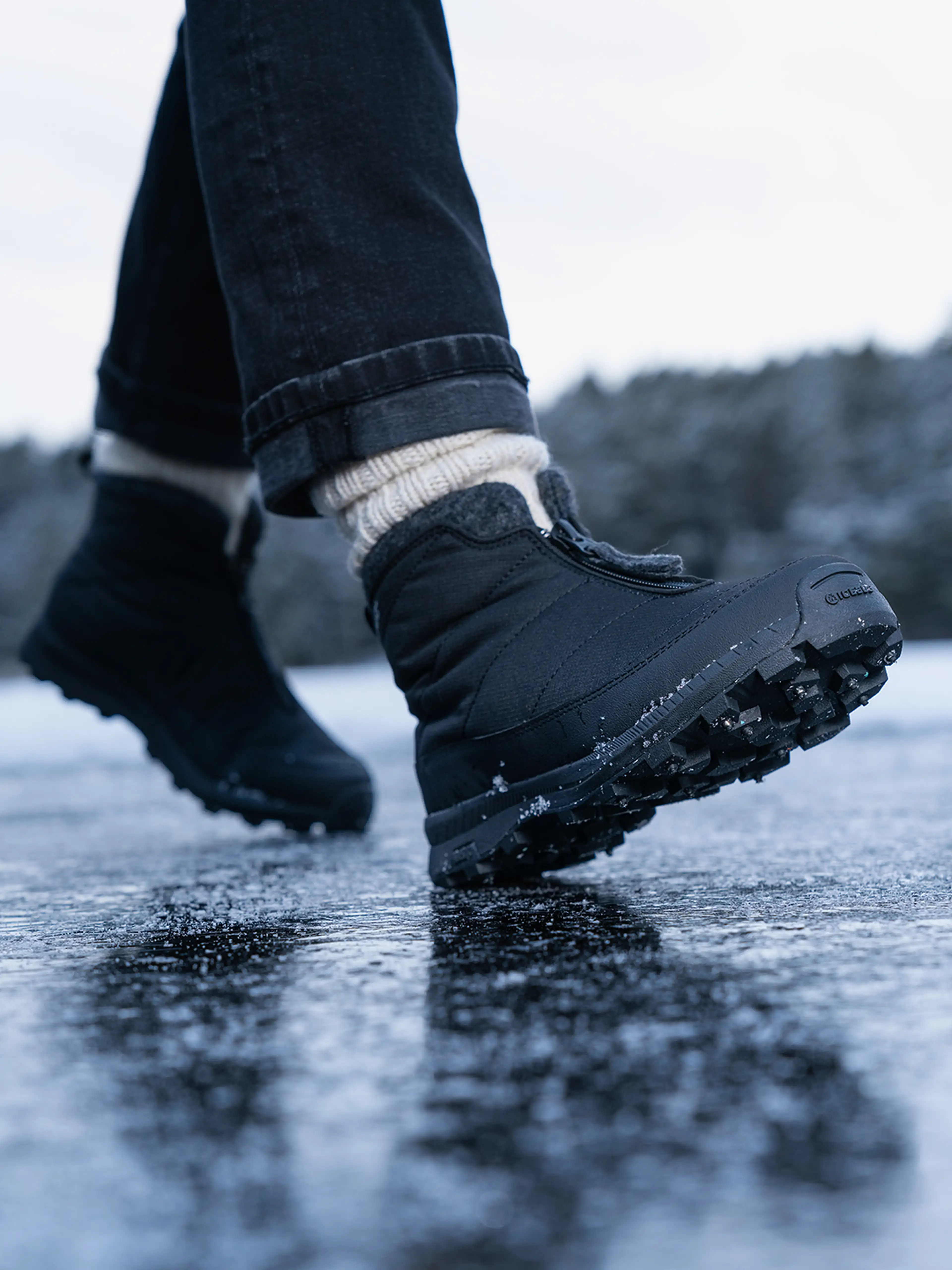 Close-up of a person wearing black winter boots and jeans walking on an icy surface, with a blurred snowy landscape in the background.