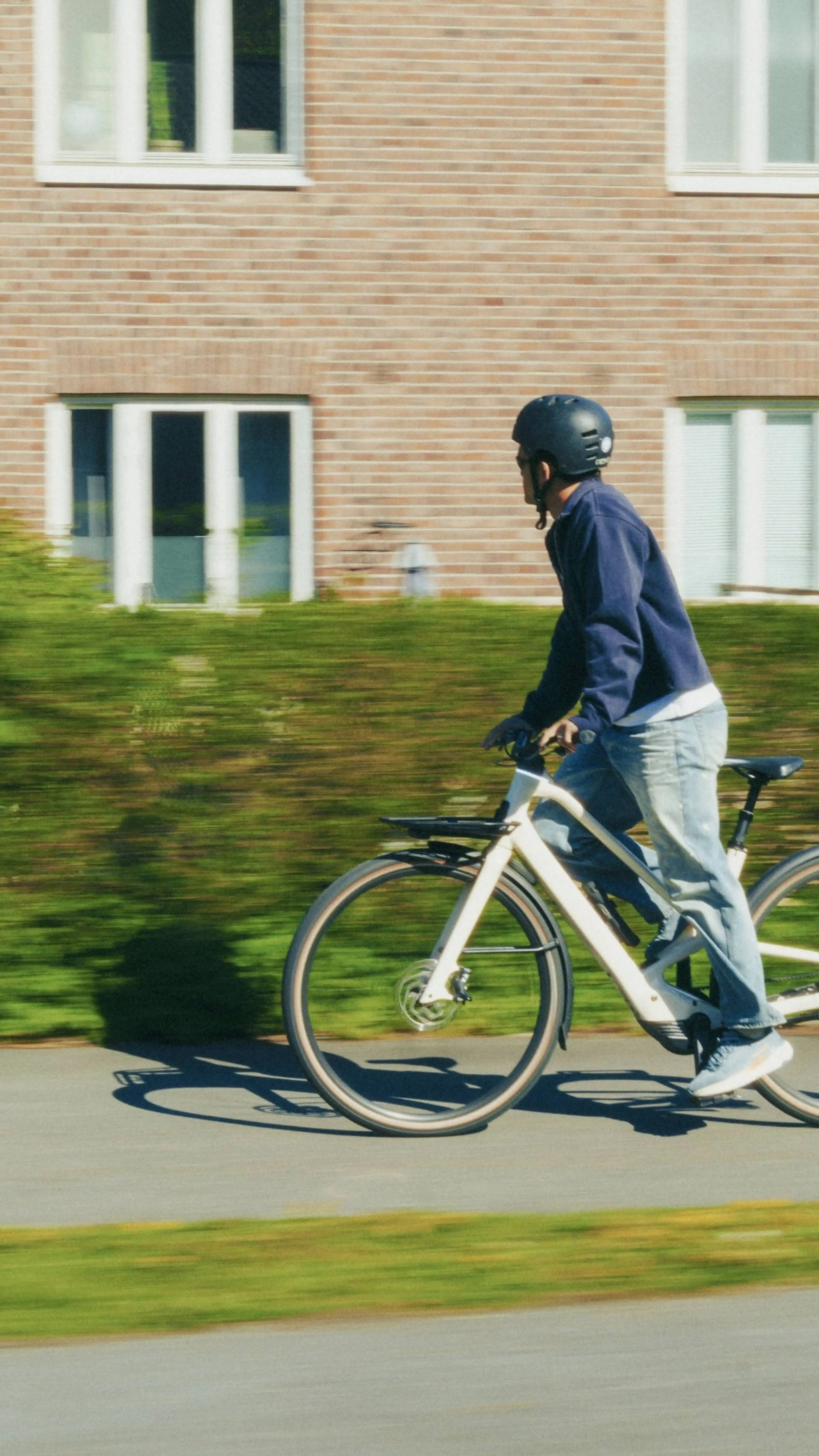 Person wearing a helmet and blue jacket rides a bicycle past a brick building on a sunny day.